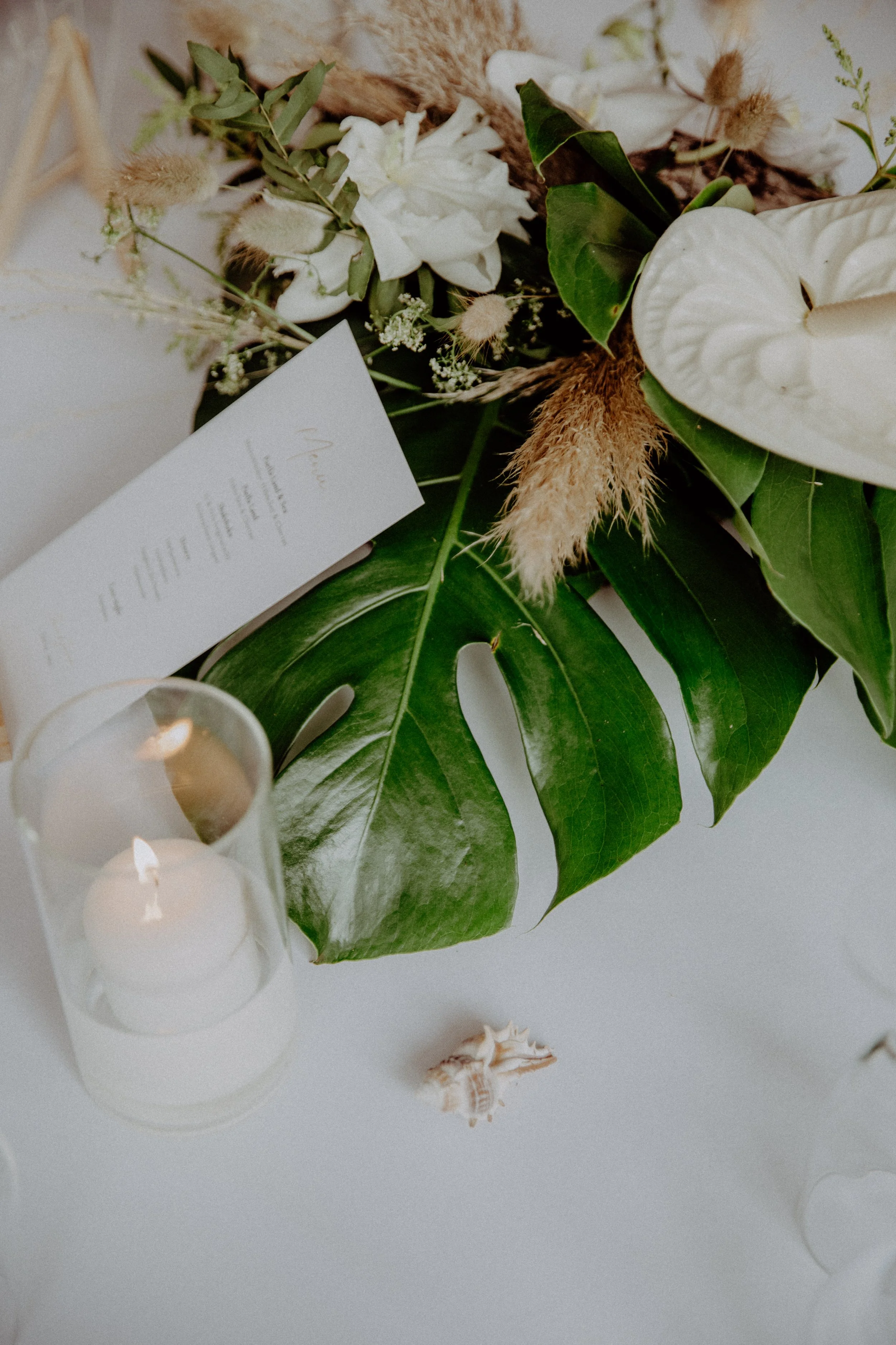 Eine flache Anordnung aus einem großen grünen Monstera-Blatt, weißen Blumen, Heidekraut, und anderen Pflanzenteilen, ergänzt durch eine Kerze in einem Glas, und eine Einladungskarte. Elemente deuten auf eine elegante, natürliche Hochzeitsdekoration.