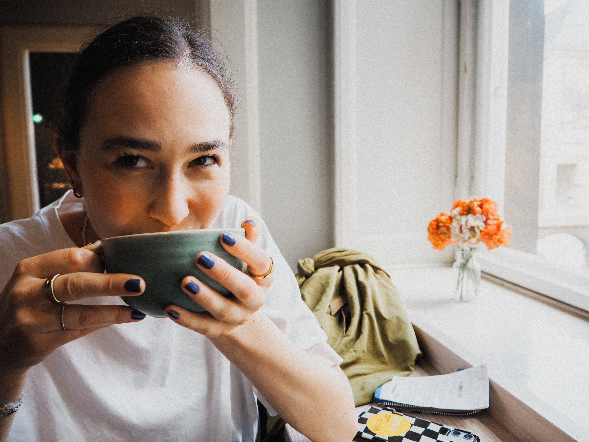 Junge Frau trinkt Kaffee in einem Café bei Tageslicht, neben ihr auf dem Tisch liegen ein Handy, eine Zeitschrift, eine Tasche und ein Blumenstrauß mit orangefarbenen Blumen auf dem Fensterbrett.