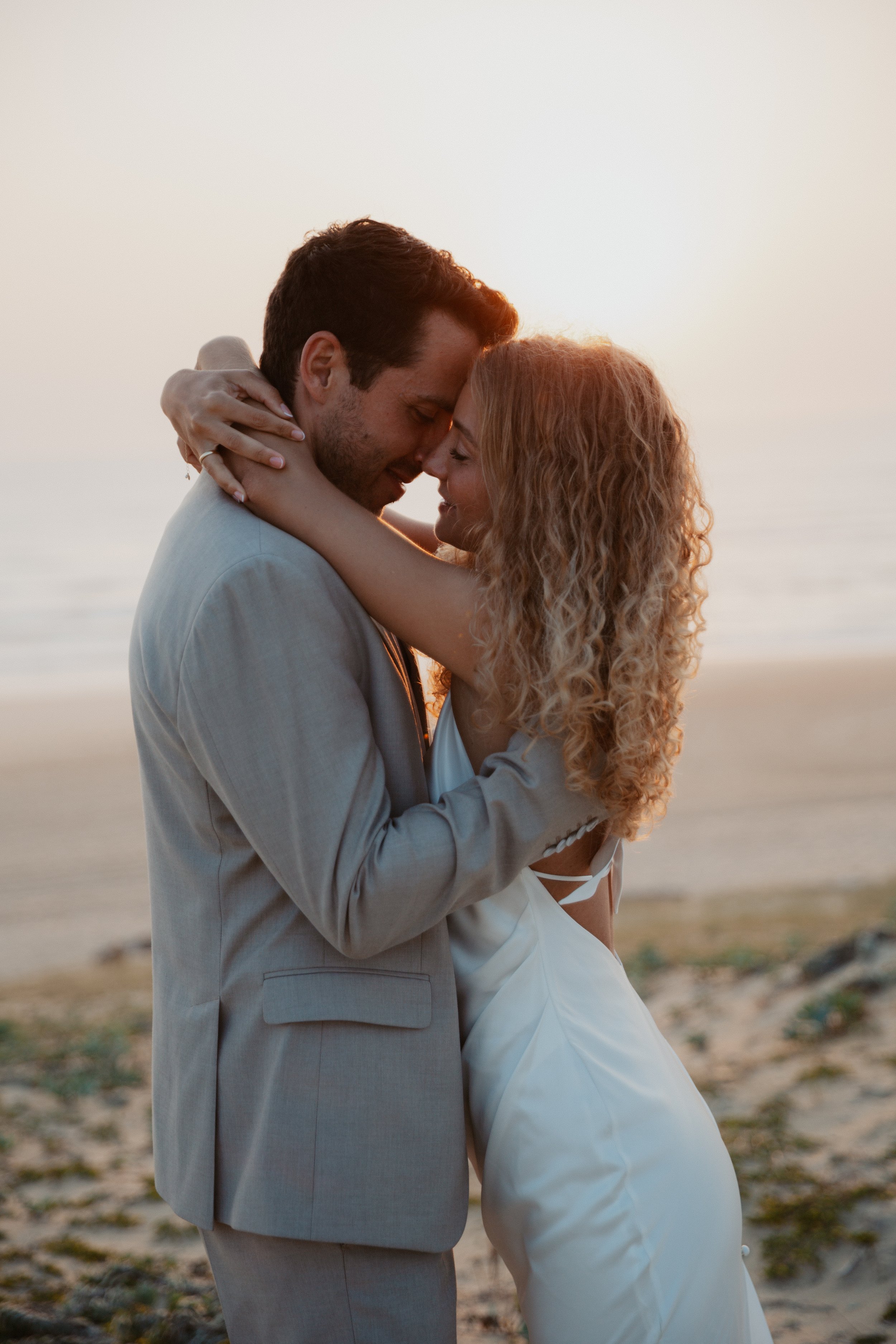 Ein verliebtes Paar umarmt sich am Strand bei Sonnenuntergang und trägt Hochzeitskleidung.