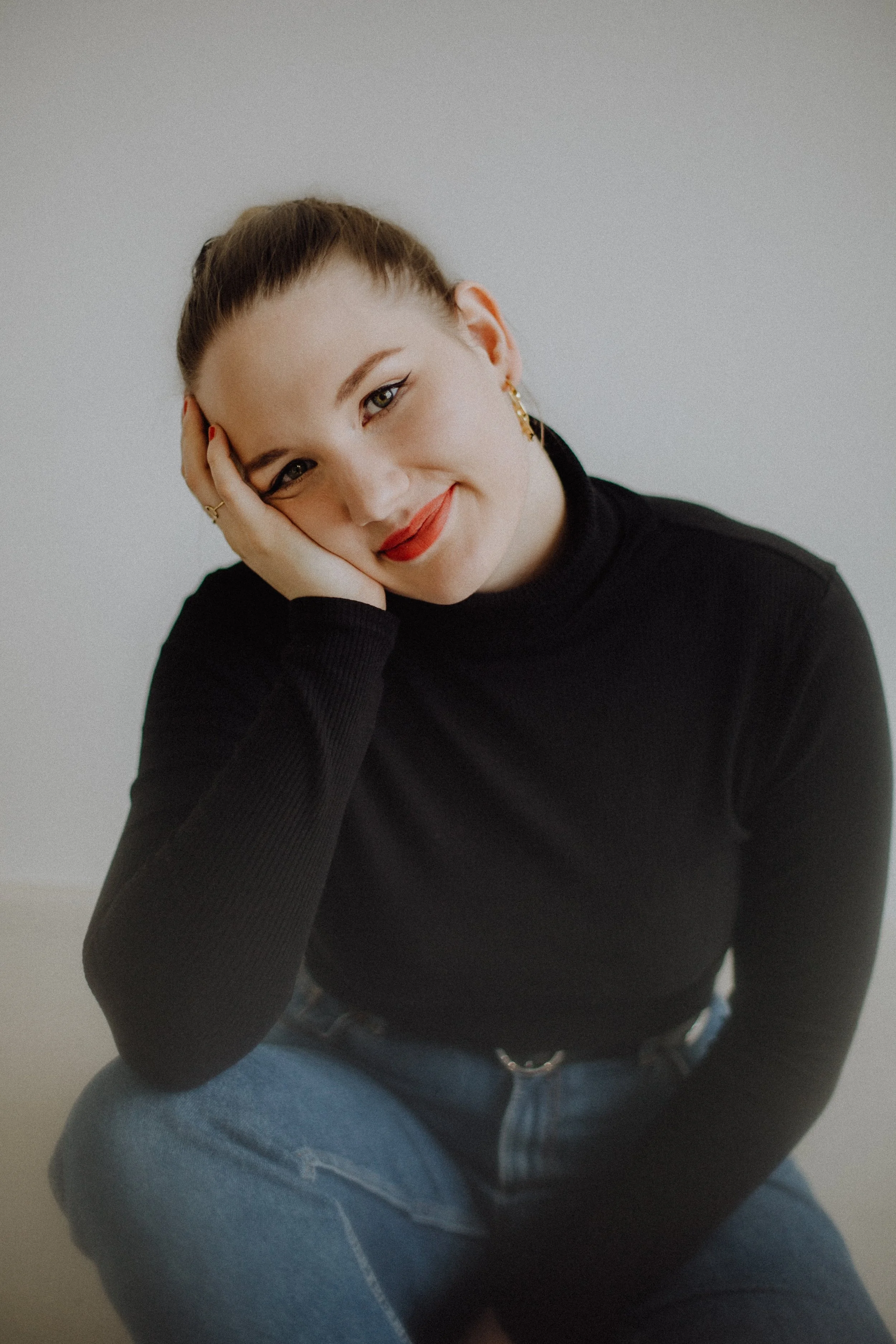 Eine junge Frau mit braunen Haaren in einem schwarzen Rollkragenpullover und blauen Jeans, sitzt lächelnd und mit einem Arm auf ihrem Knie, vor hellem Hintergrund.