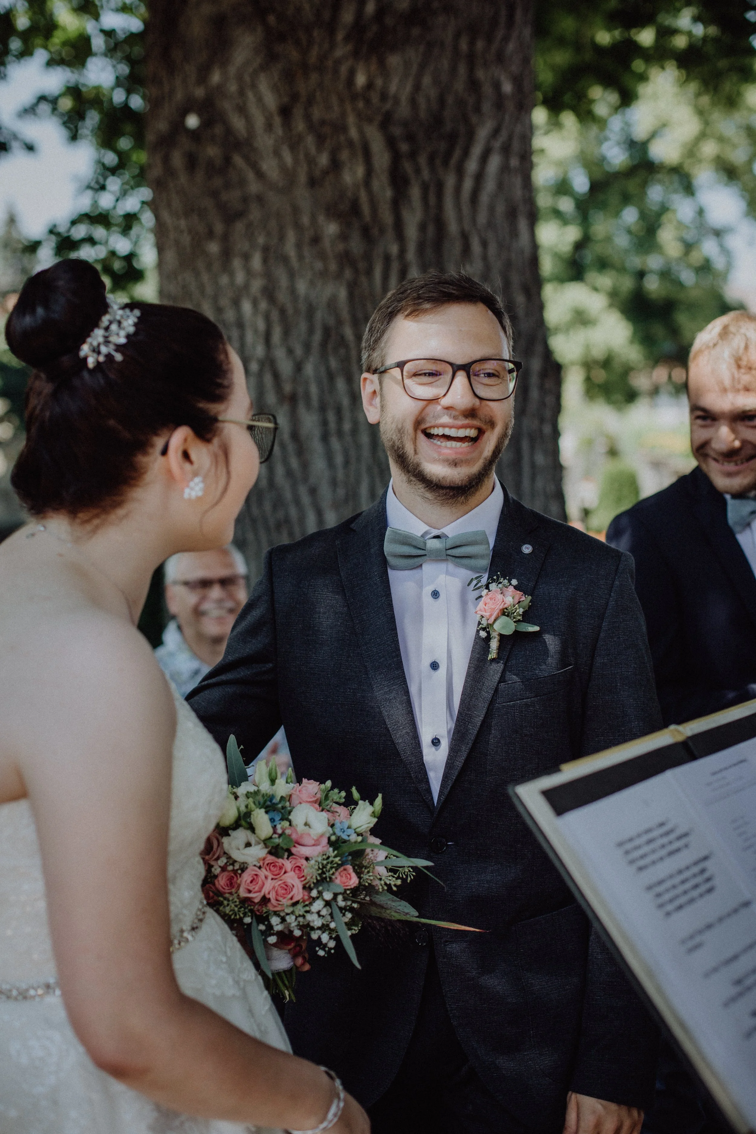Ein Brautpaar bei ihrer Hochzeit, lachend und sich in die Augen blickend, im Freien unter einem Baum mit Freunden im Hintergrund.