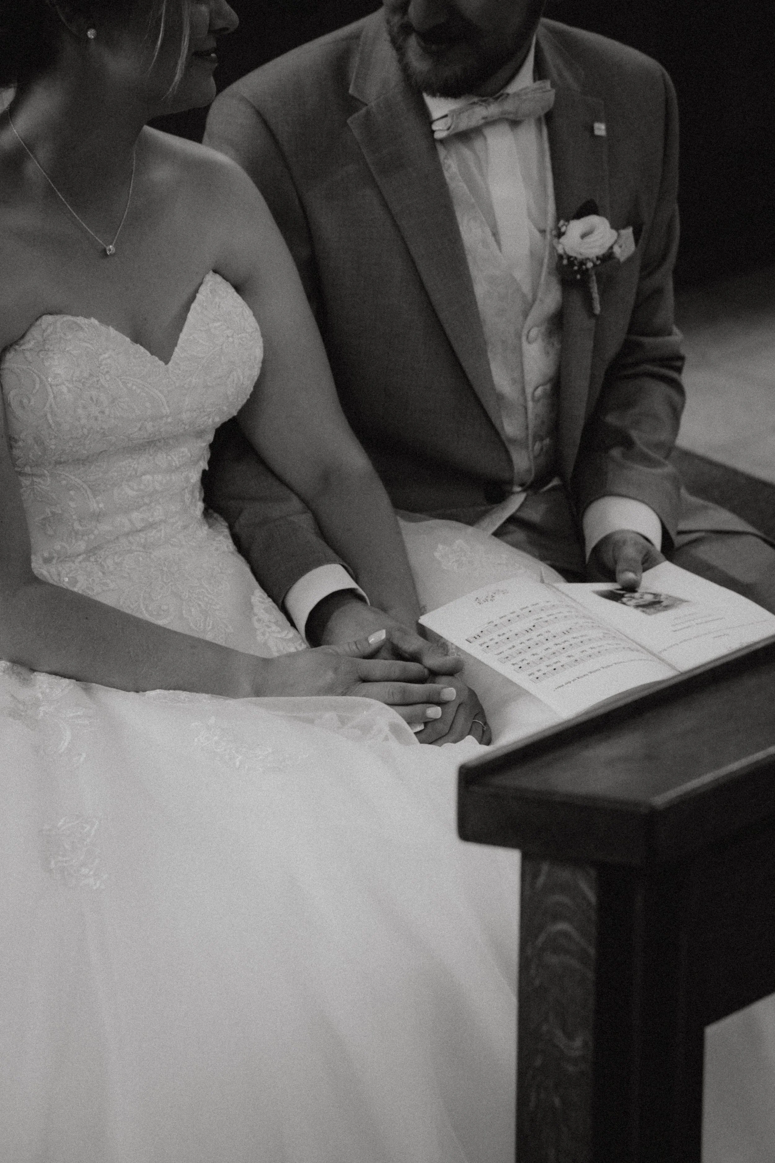 Hochzeitspaar sitzt zusammen, das Brautpaar hält die Hände, das Brautkleid ist detailliert und der Mann trägt einen Anzug mit Fliege, beide schauen auf ein Buch oder Heft.