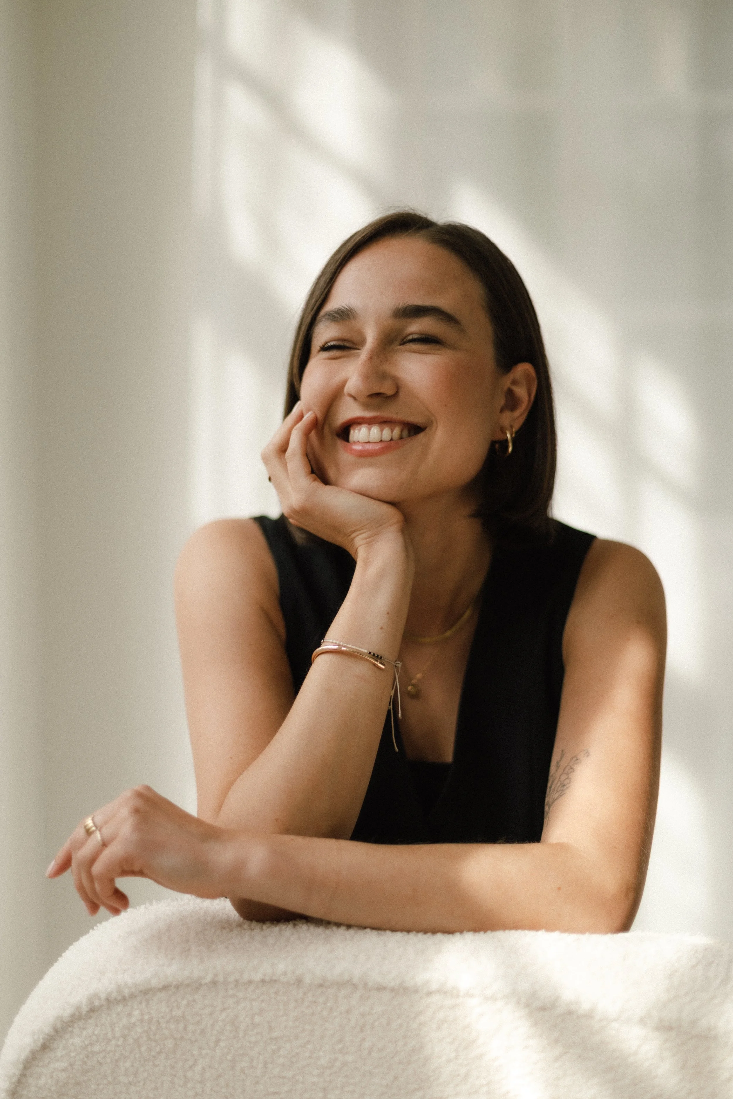 Lächelnde junge Frau mit dunklen, schulterlangen Haaren, trägt schwarze Kleidung, goldene Ohrringe, Halsketten und Armreifen, sitzt auf einem weißen Möbelstück vor einem hellen Hintergrund mit Sonnenlicht.