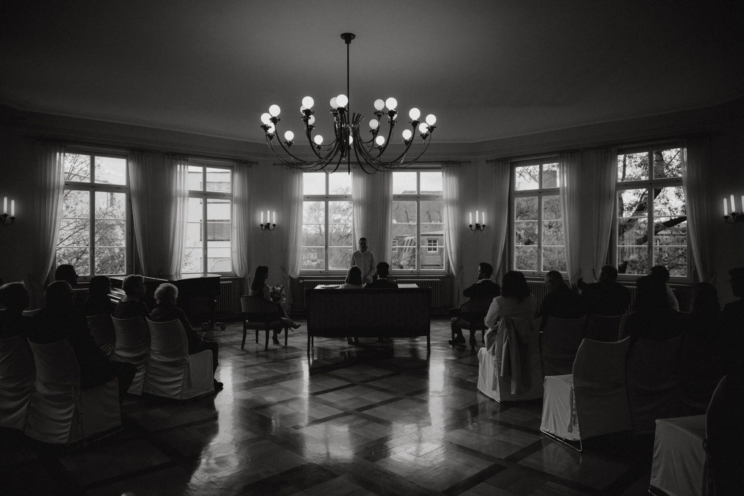 Schwarz-weiß-Foto einer formellen Versammlung in einem eleganten Raum mit großen Fenstern, einem Kronleuchter und mehreren anwesenden Personen, die einer Rede zuhören.