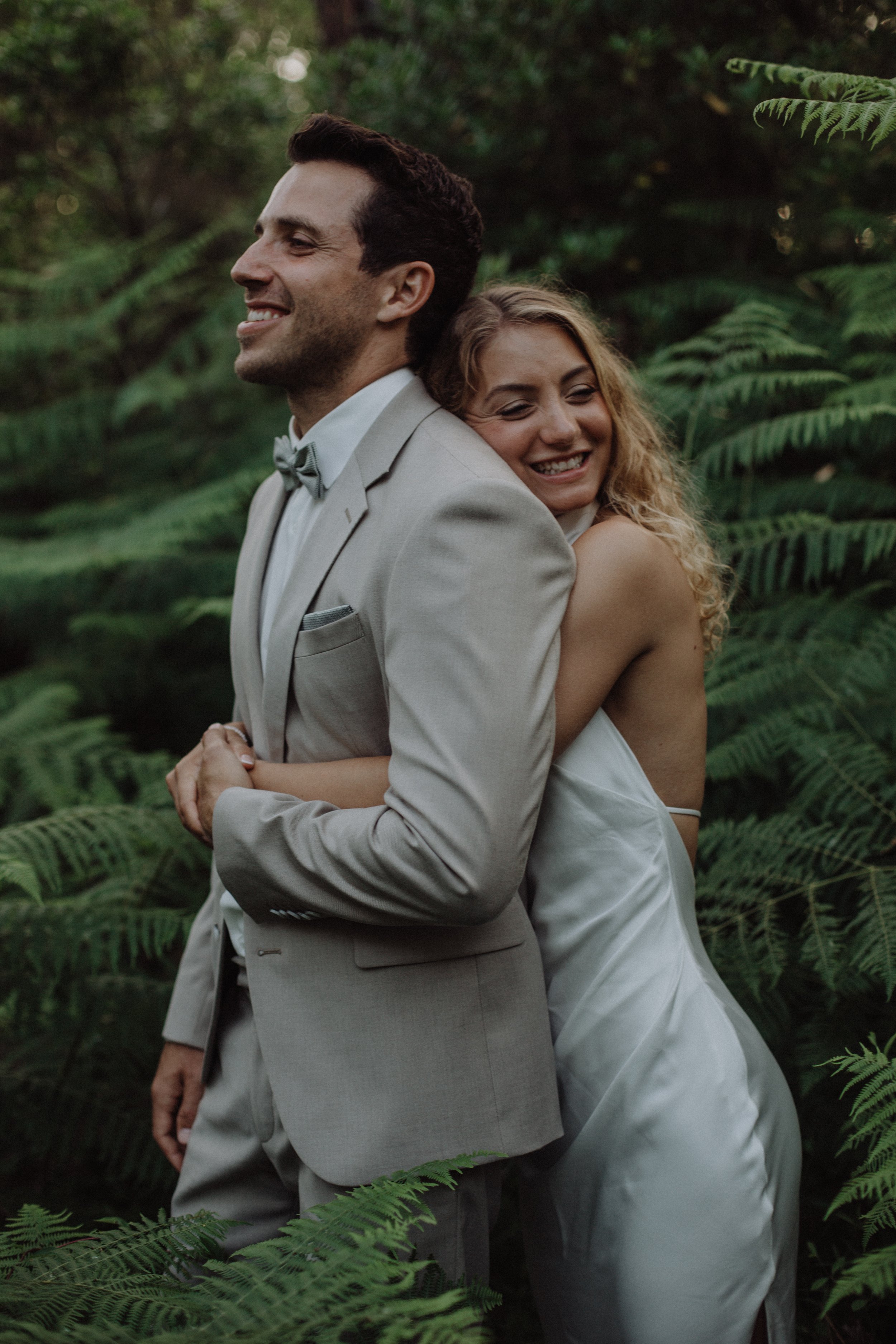 Ein glückliches Paar in formeller Kleidung, umarmt sich in einem grünen, bewaldeten Ausgang, wahrscheinlich bei einer Hochzeit.