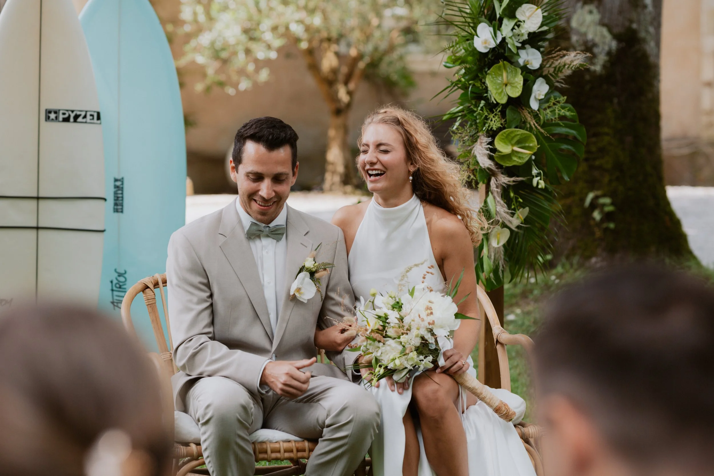 Ein lachendes Brautpaar bei einer Hochzeit im Freien, sitzend auf einem Rattanstuhl. Der Bräutigam trägt einen hellgrauen Anzug mit Fliege, die Braut trägt ein weißes Hochzeitskleid und hält einen Blumenstrauß. Im Hintergrund sind grüne Pflanzen und 