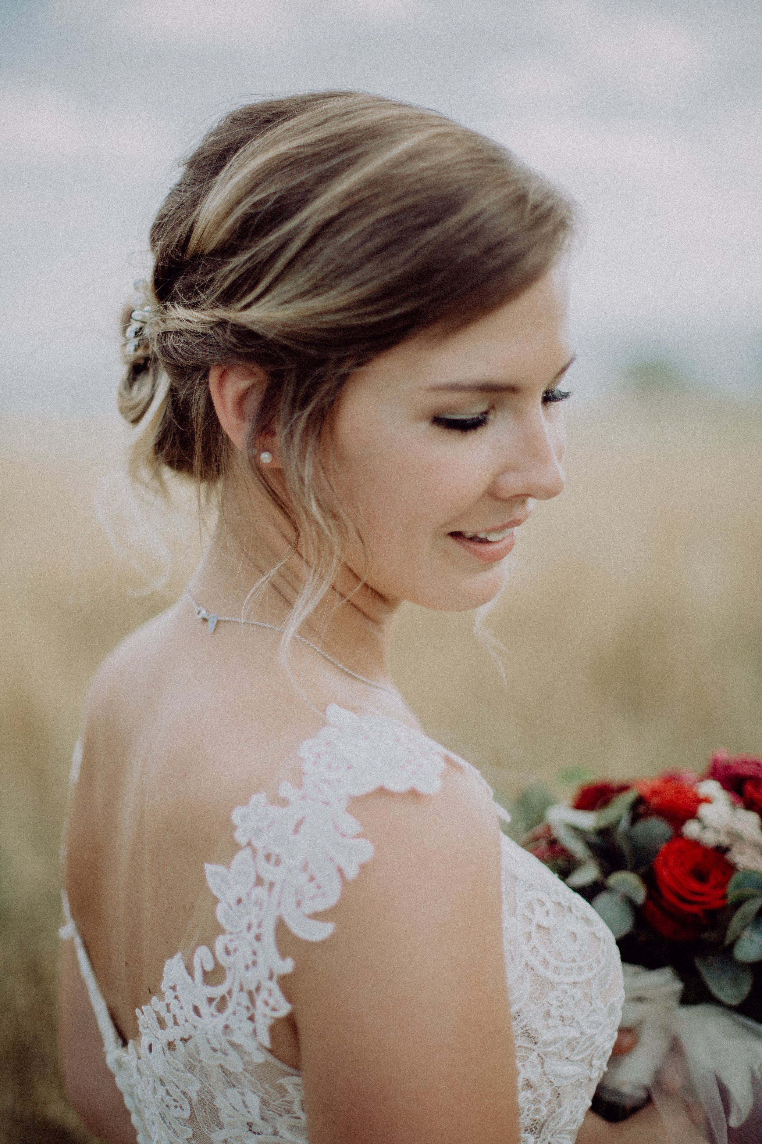 Junge Braut mit braunen, gewellten Haaren in Hochzeitskleid hält einen Blumenstrauß im Freien.