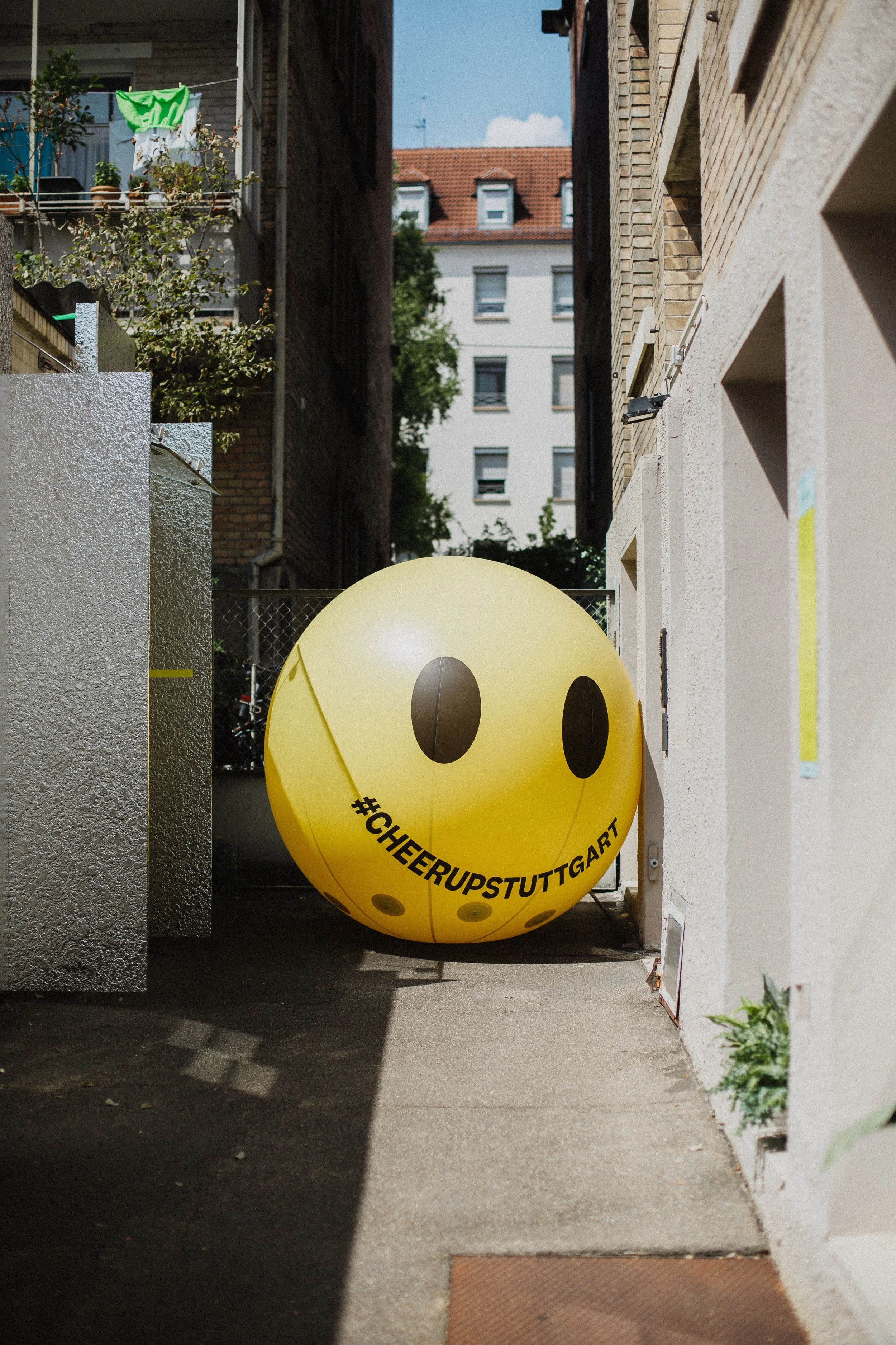 Gelber Ball mit Smile-Gesicht und dem Hashtag #CHEERUPSTUTTGART in einer engen Gasse zwischen Gebäuden, im Hintergrund ein mehrstöckiges Haus.