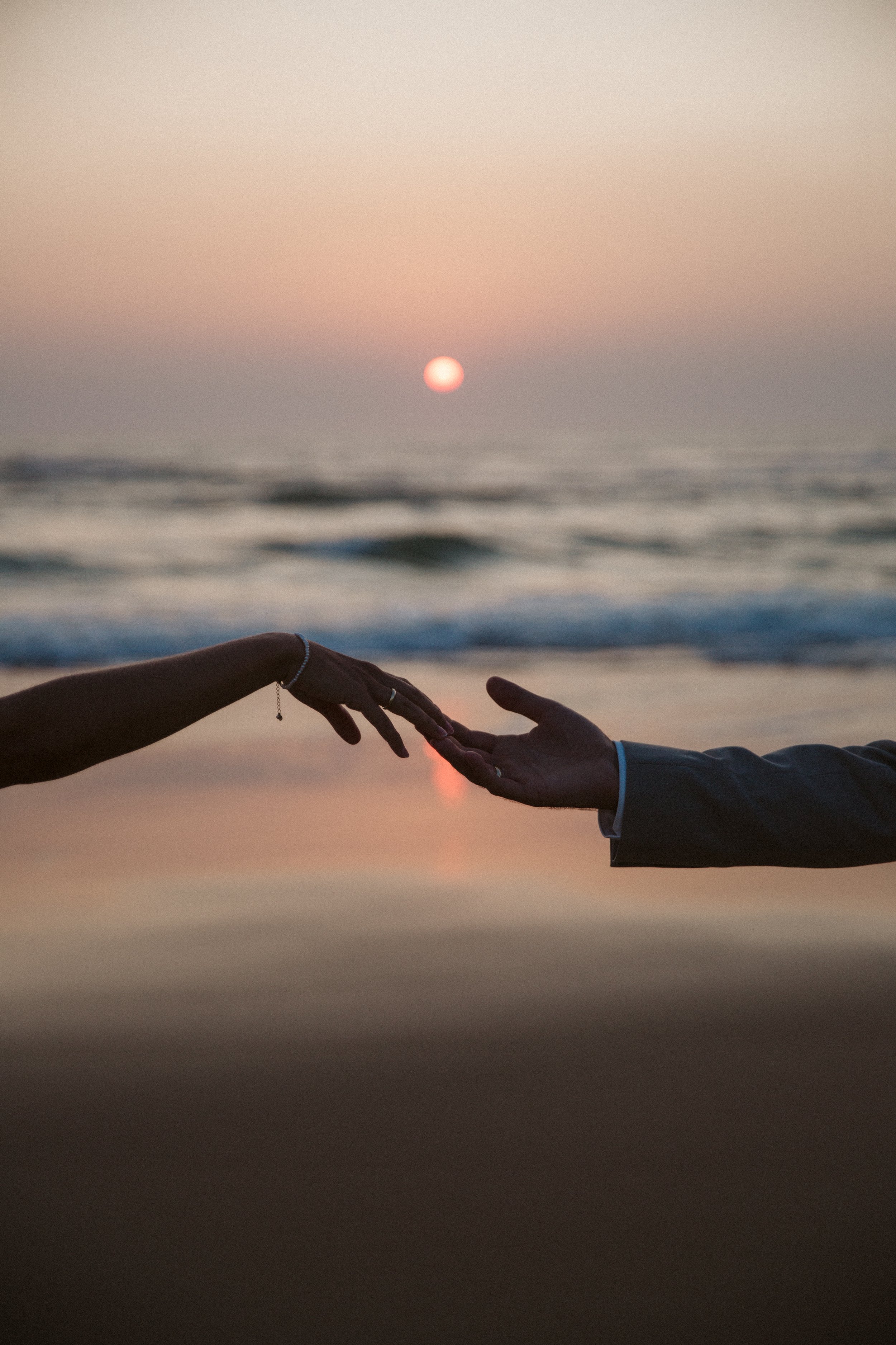 Zwei Menschen berühren sich mit den Händen am Strand bei Sonnenuntergang, im Hintergrund das Meer und die untergehende Sonne.