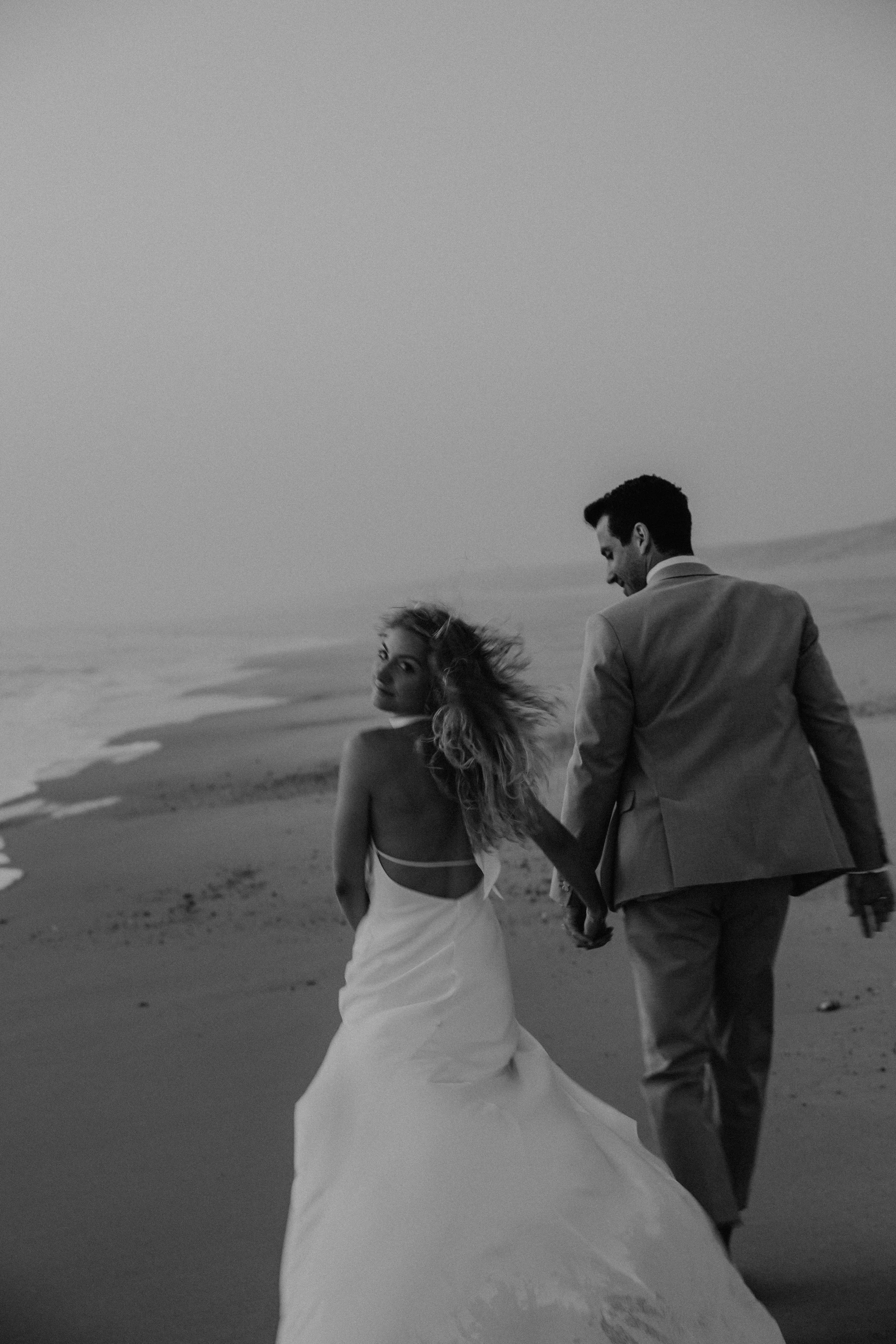 Ein verliebtes Paar in formeller Kleidung, das am Strand bei Sonnenuntergang Hand in Hand geht, schwarz-weiß Aufnahme. Die Frau trägt ein weißes Hochzeitskleid und der Mann einen beigen Hochzeitsanzug.