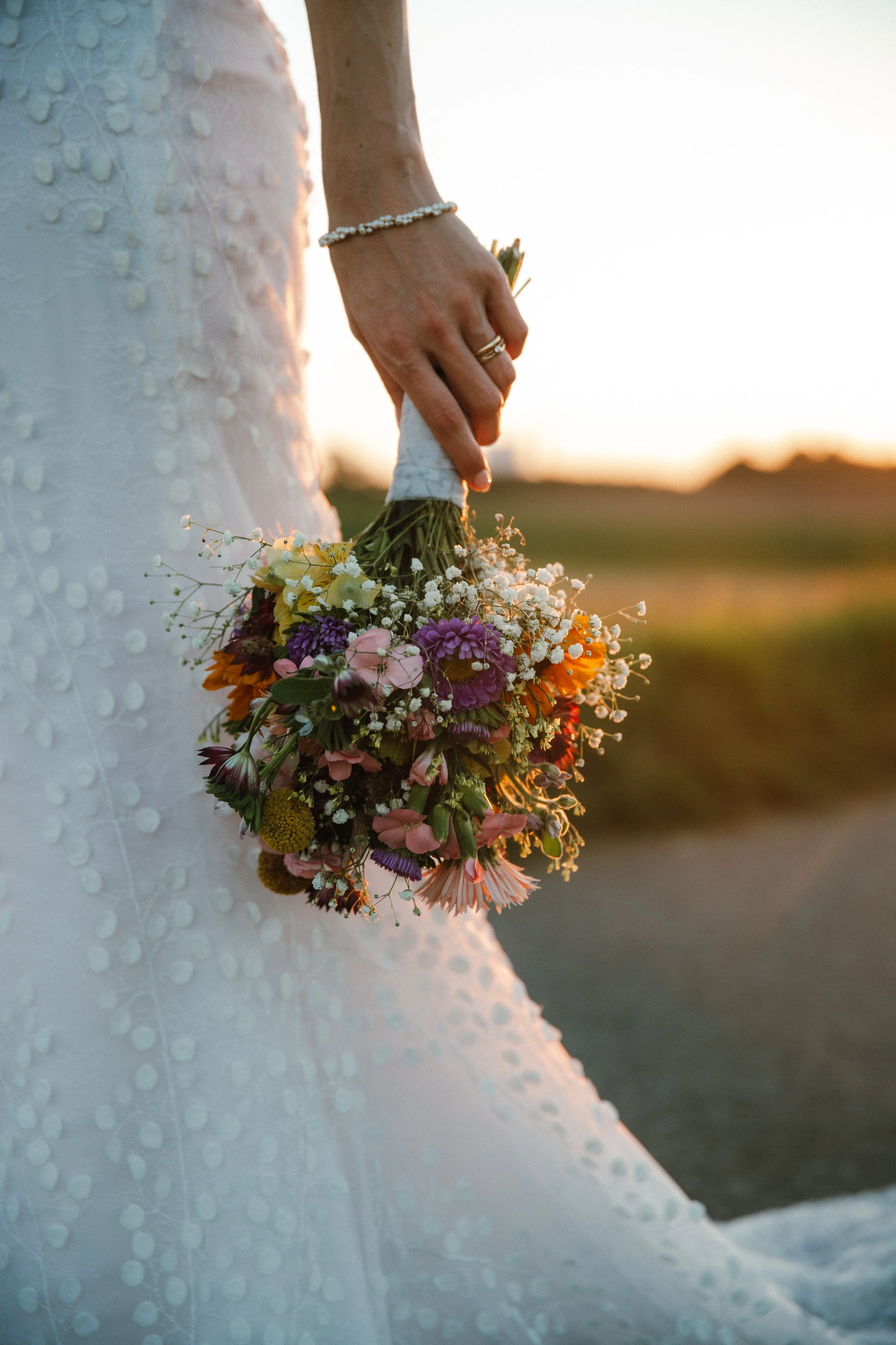 Nahaufnahme einer Hand, die einen kleinen bunten Blumenstrauß bei Sonnenuntergang hält, mit einem weißen Kleid im Hintergrund.