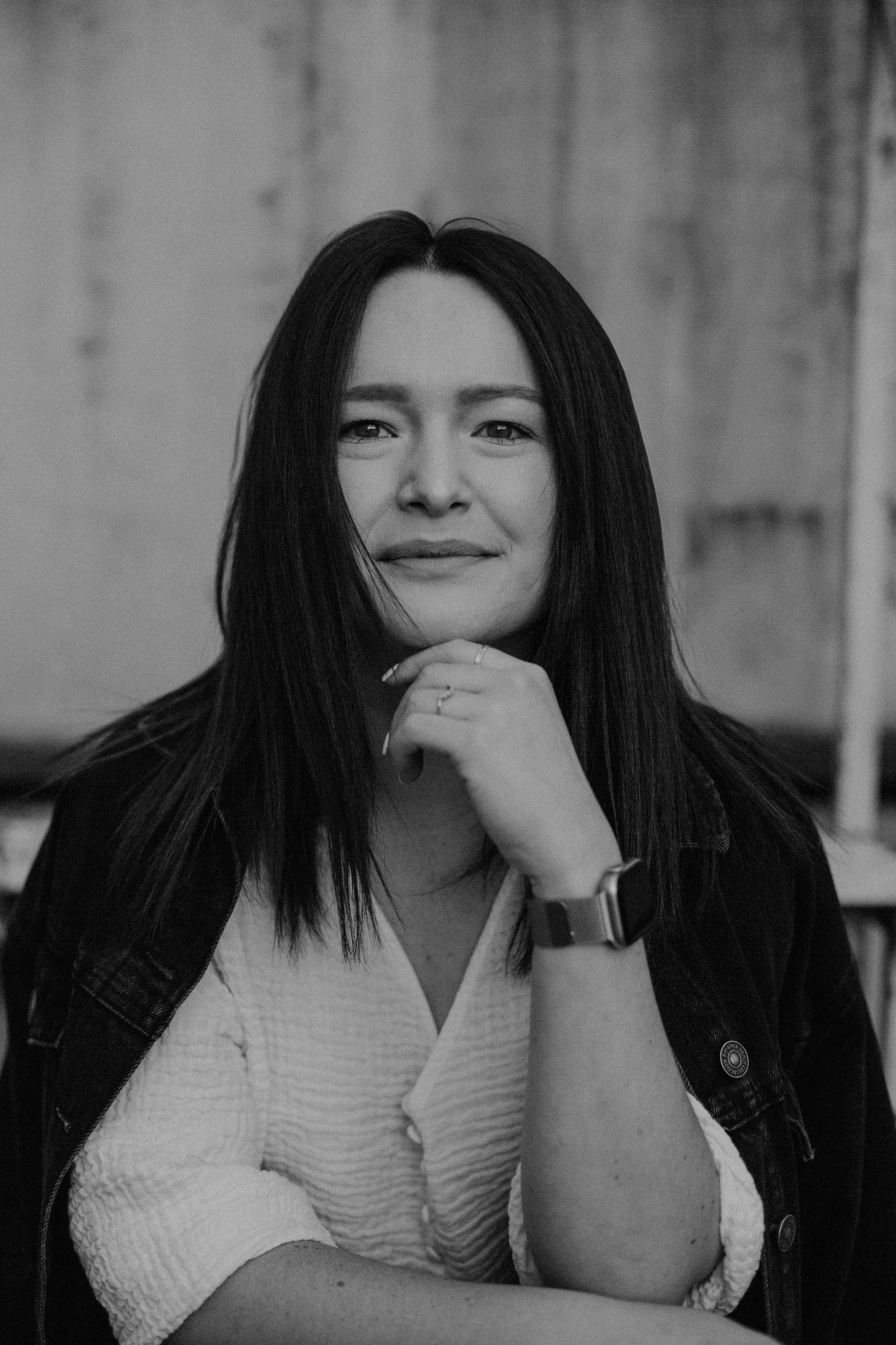 Schwarz-weiß Portrait einer Frau mit langem dunklem Haar, die nachdenklich mit der Hand am Kinn vor einer unscharfen urbanen Kulisse schaut.