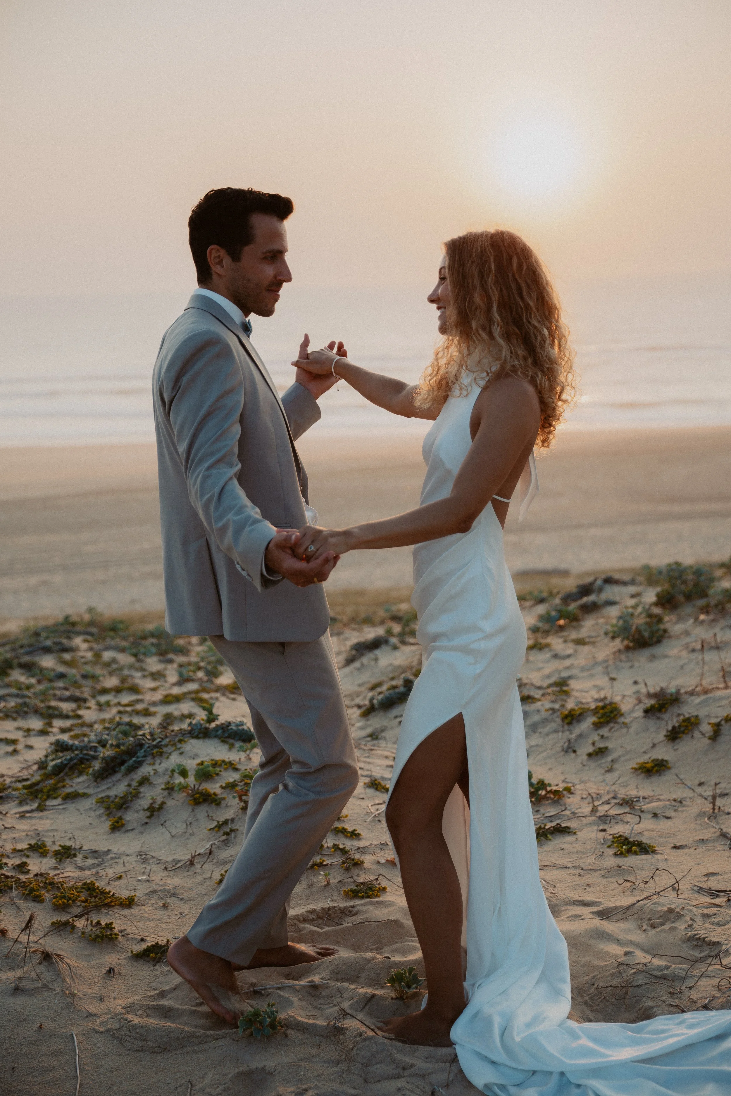 Ein Paar heiratet am Strand bei Sonnenuntergang, die Frau trägt ein weißes Kleid mit hohen Beinen, der Mann einen grauen Anzug, sie tanzen und halten Händchen.
