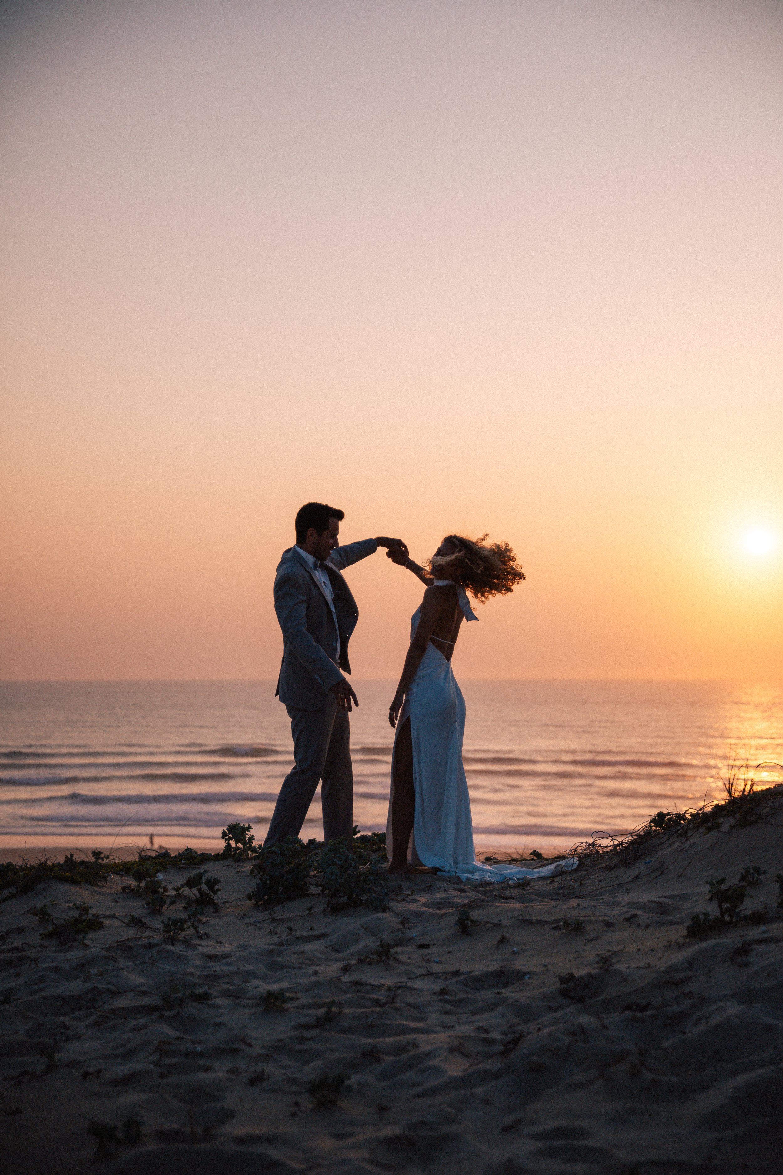 Ein Paar tanzt am Strand bei Sonnenuntergang, der Mann hält die Hand der Frau, beide sind in formeller Kleidung. Der Himmel ist orange und pink, das Meer ist ruhig.