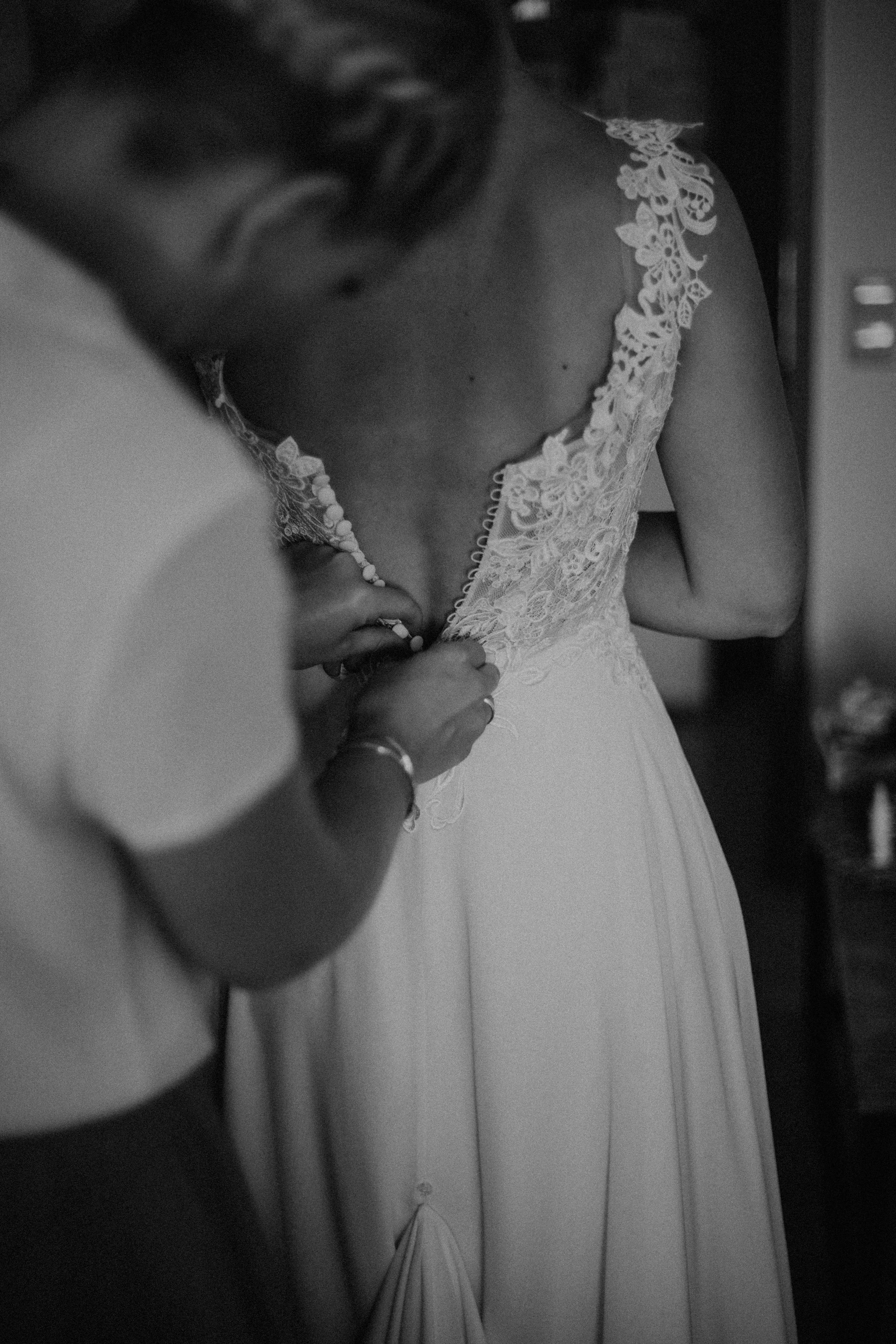 Schwarz-Weiß-Foto einer Braut, die bei ihrer Hochzeit in einem eleganten Kleid mit Stickerei am Oberteil ist, während eine Frau ihr den Verschluss des Kleides schließt.