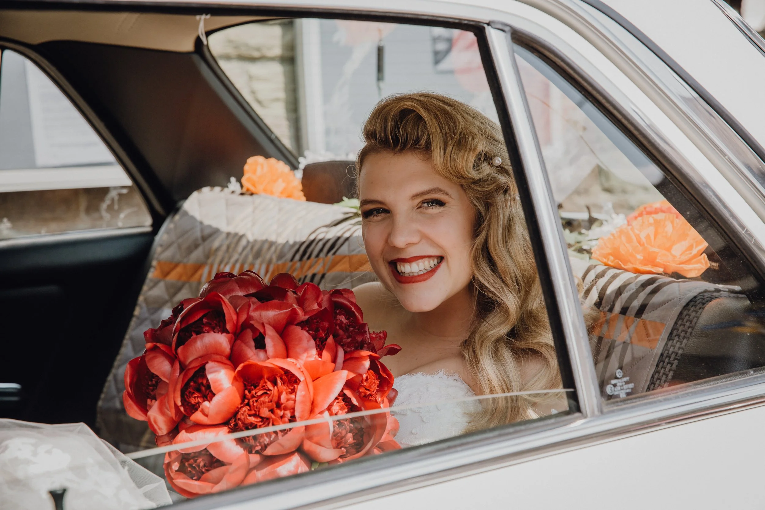 Junge Frau sitzt in einem Auto, trägt ein weißes Kleid, hält einen großen roten Blumenstrauß, lacht breit und zeigt Freude, mit weiteren Blumen im Auto.