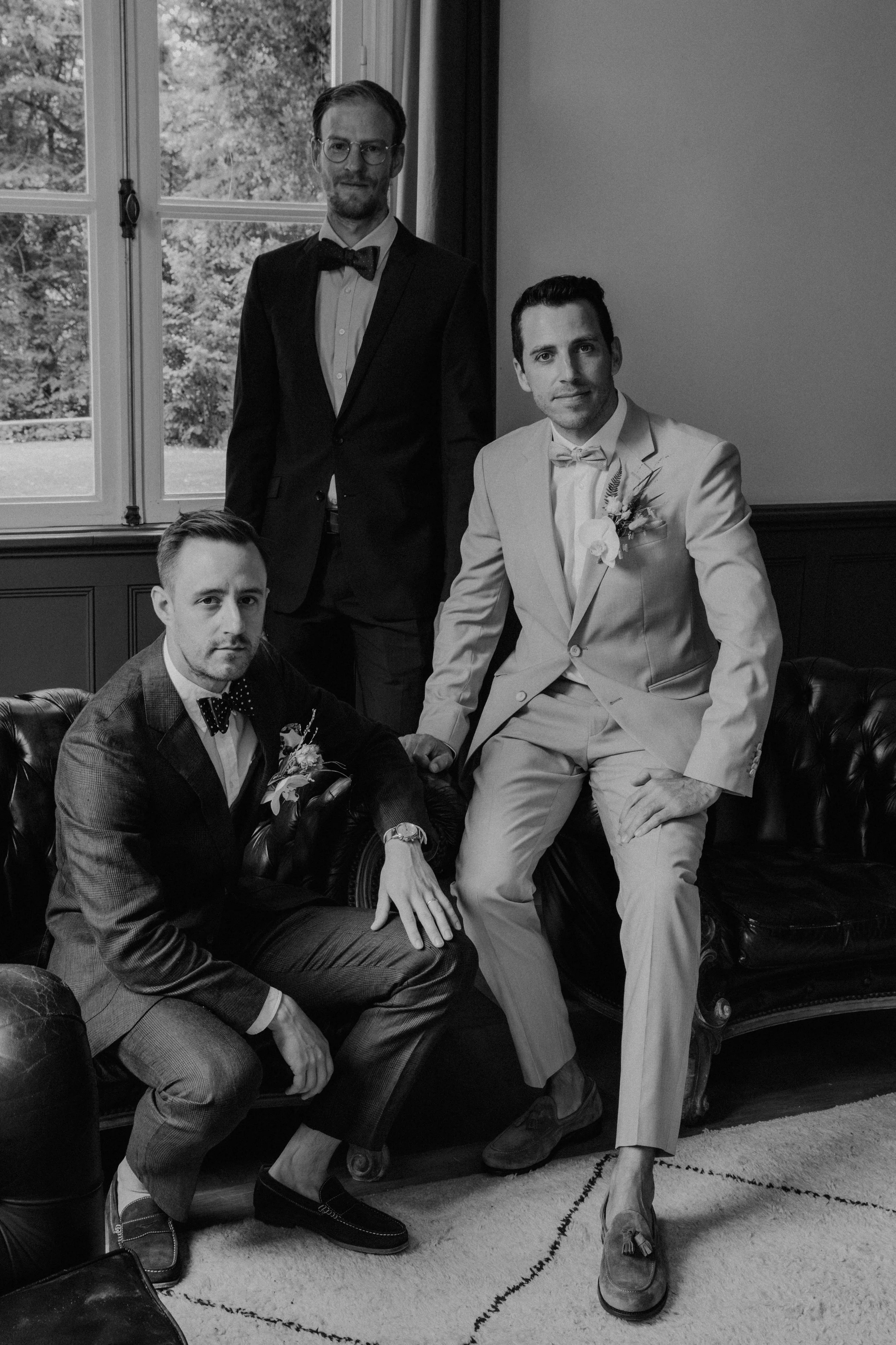 Schwarz-weiß-Foto von vier Männern bei einer formellen Veranstaltung, wahrscheinlich eine Hochzeit, im Wohnzimmer vor einem Fenster mit Blick auf Bäume. Einer sitzt auf einer Couch, ein anderer sitzt darauf, der dritte steht hinter ihnen, alle in ele