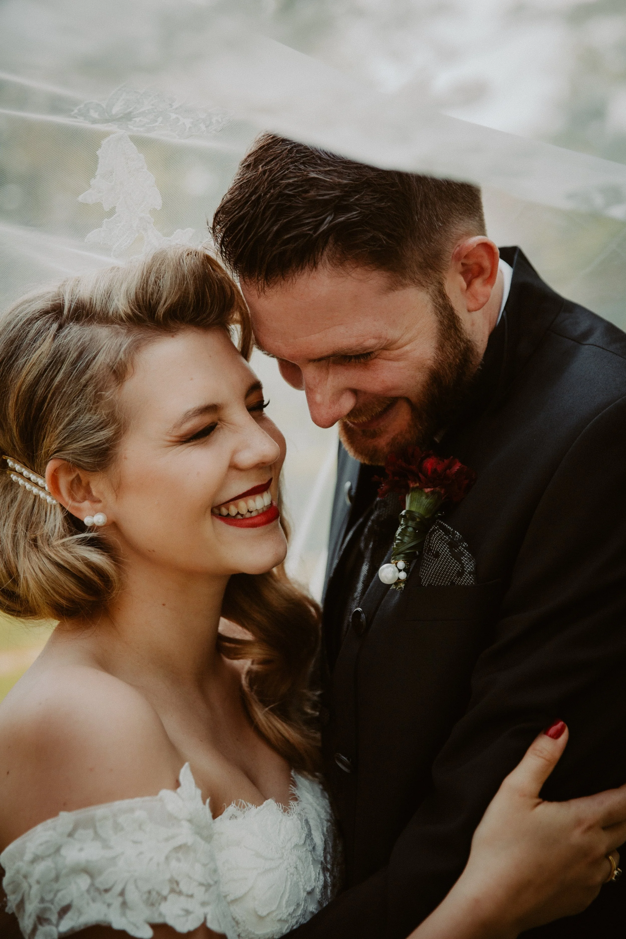 Ein glückliches Brautpaar lächelt sich an, umarmt sich und trägt formelle Kleidung, auf einer Hochzeit. Die Braut hat lockige, braune Haare, rote Lippen und trägt Perlenohrringe, während der Bräutigam einen dunklen Anzug trägt.