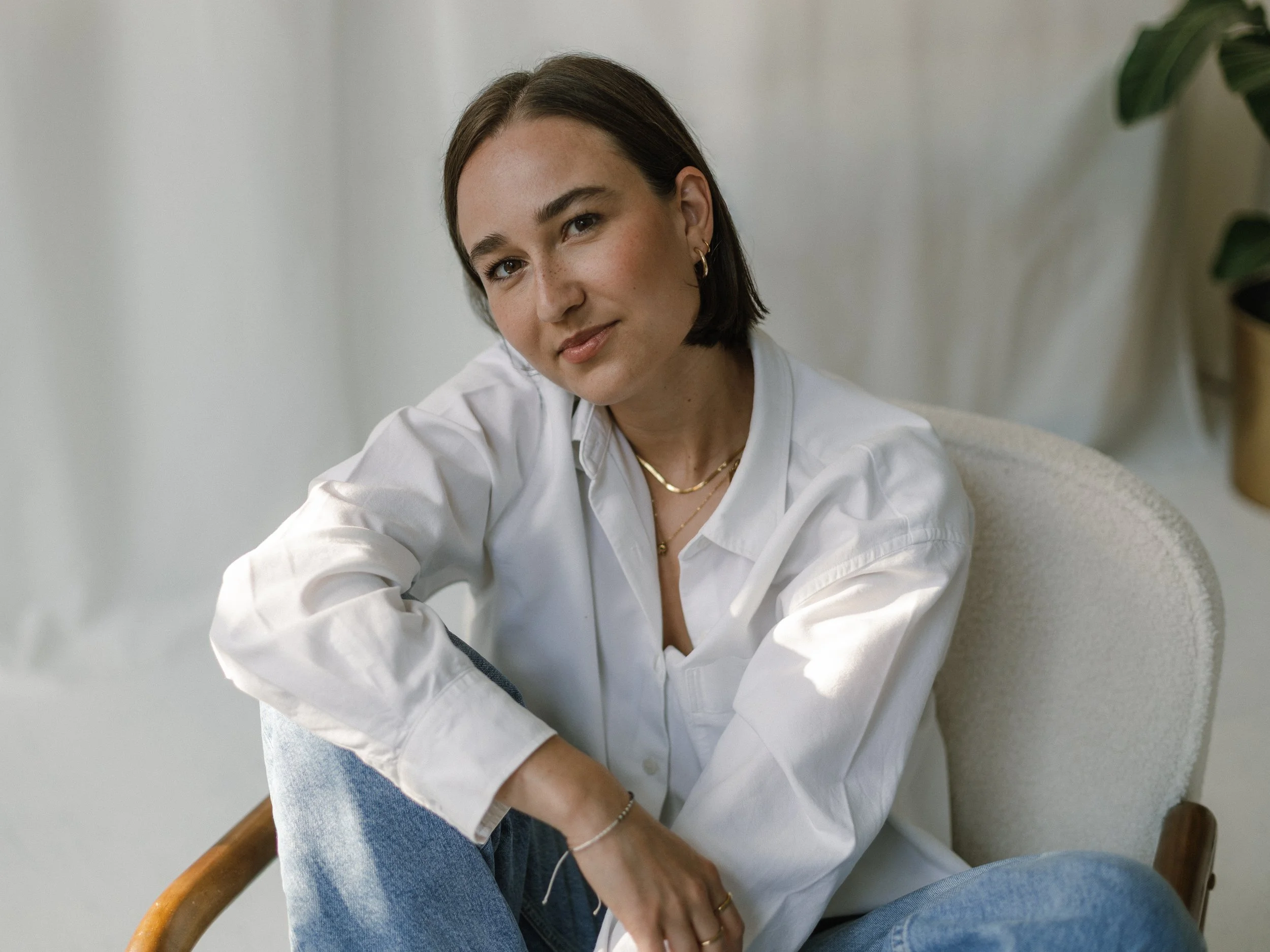 Eine junge Frau sitzt entspannt in einem weißen Hemd und blauen Jeans in einem hellen Raum, umgeben von Pflanzen.