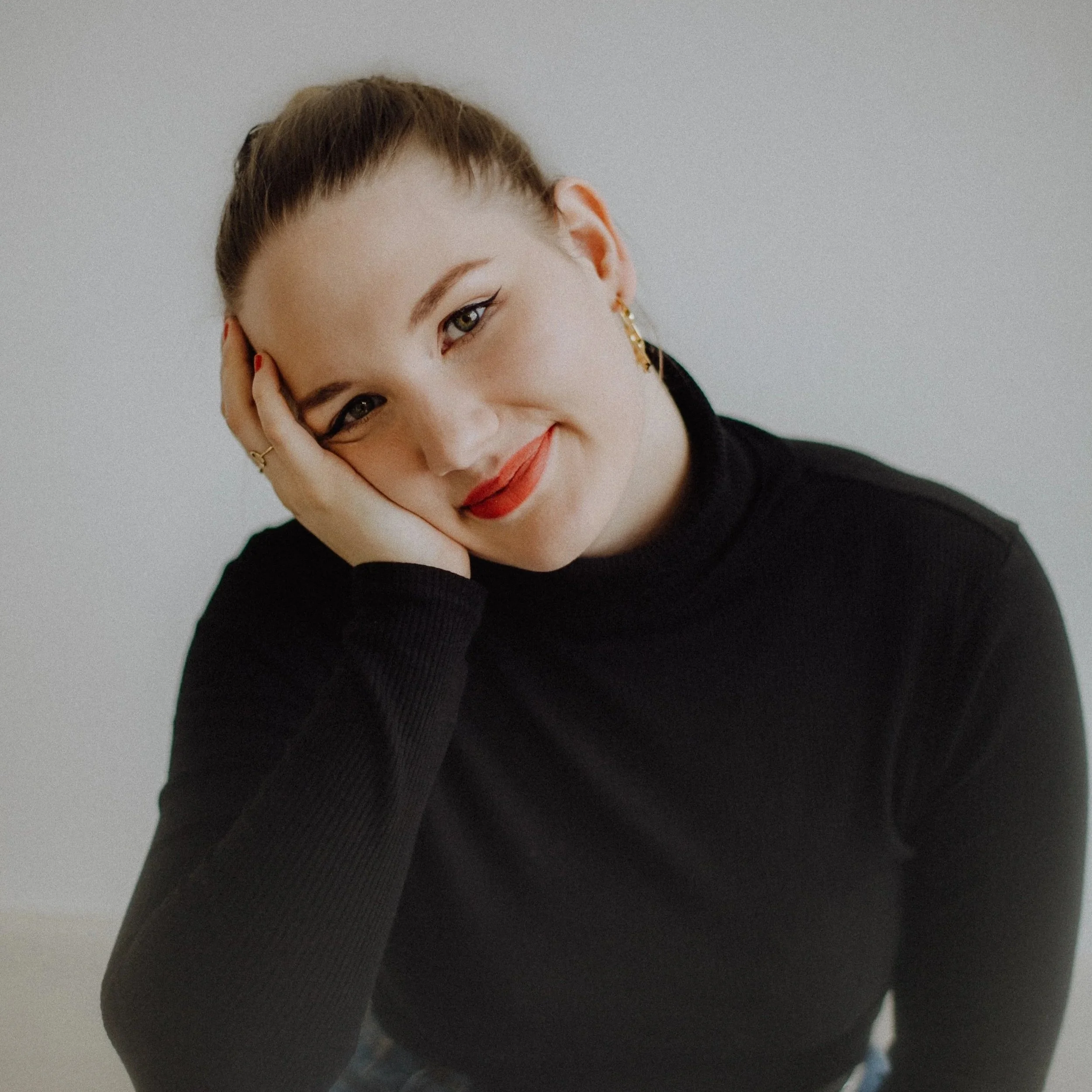 Nahaufnahme eines lächelnden jungen Mädchens mit braunen Haaren, schwarzem Turtleneck und roten Lippen, die auf einem weißen Hintergrund sitzt.