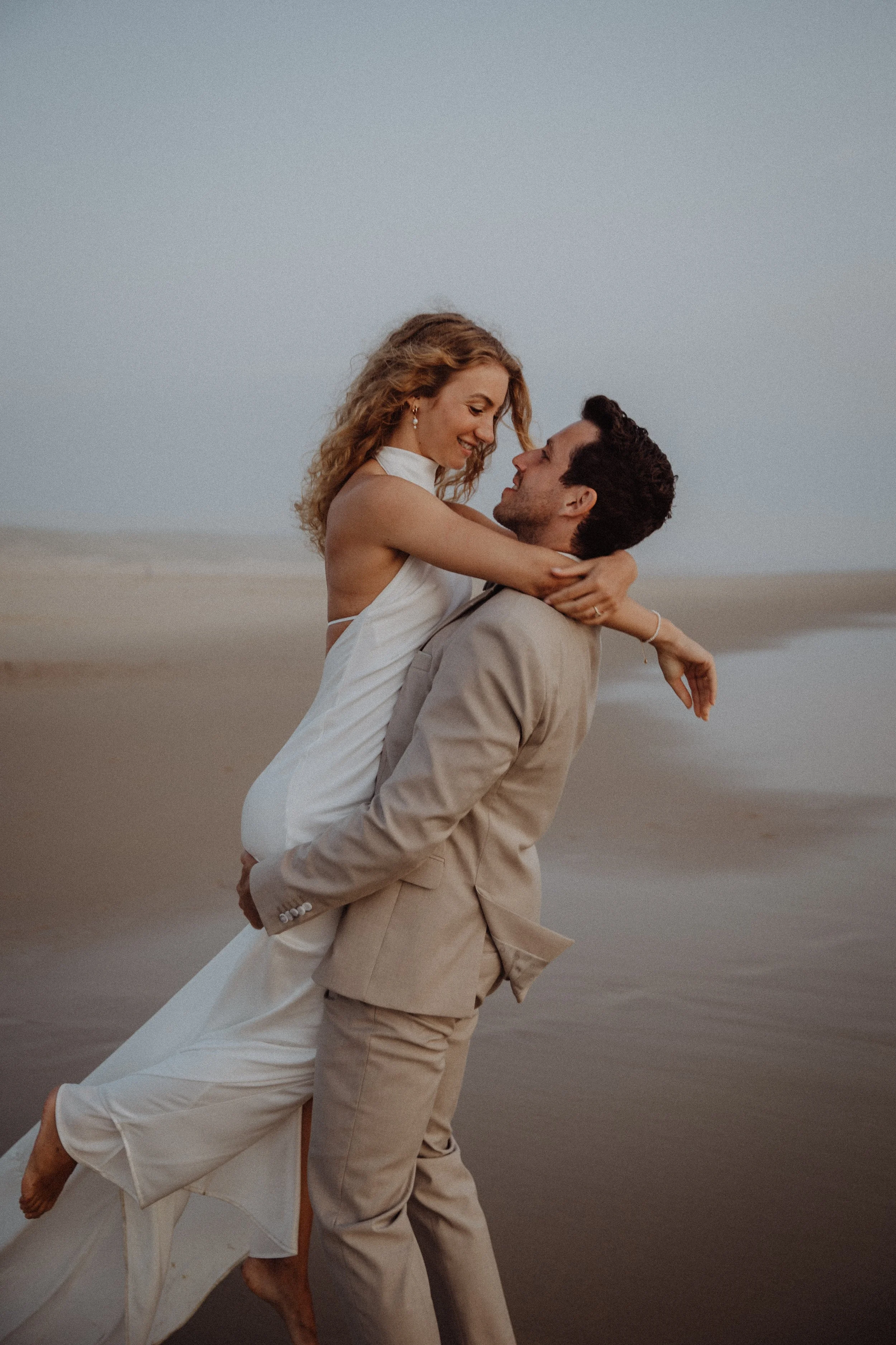 Ein verliebtes Paar, der Mann trägt einen hellen Anzug, die Frau ein langes weißes Kleid, bei Sonnenuntergang am Strand.