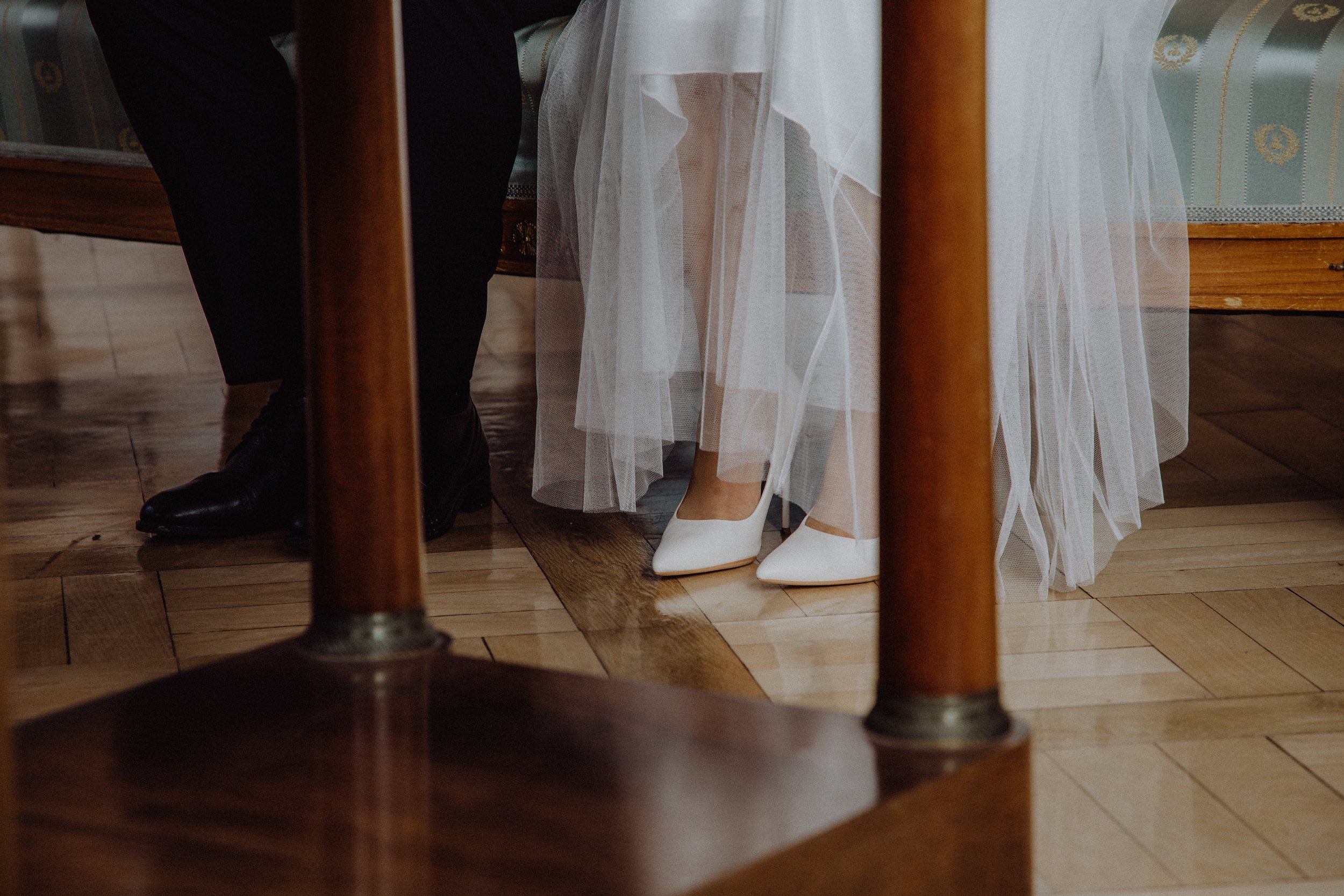 Nahaufnahme der Füße einer Hochzeitsbraut in weißen High Heels und eines Bräutigams in schwarzen Schuhen, die unter einem Tischtuch hervorschauen.