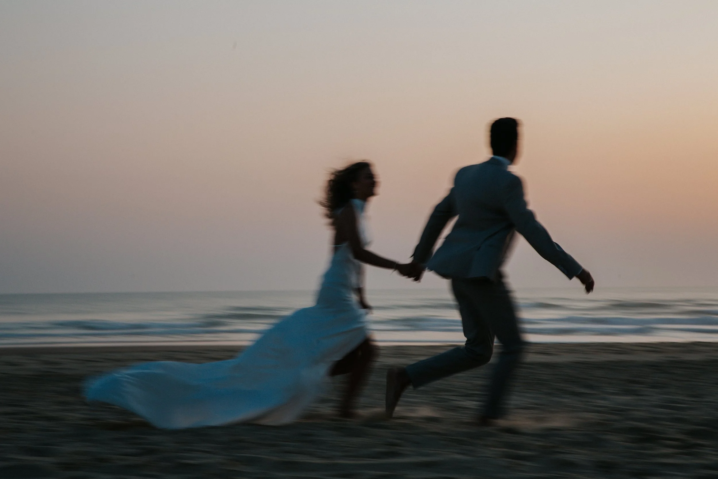 Ein Paar läuft am Strand bei Sonnenuntergang, die Frau trägt ein weißes Kleid, der Mann trägt einen Anzug.