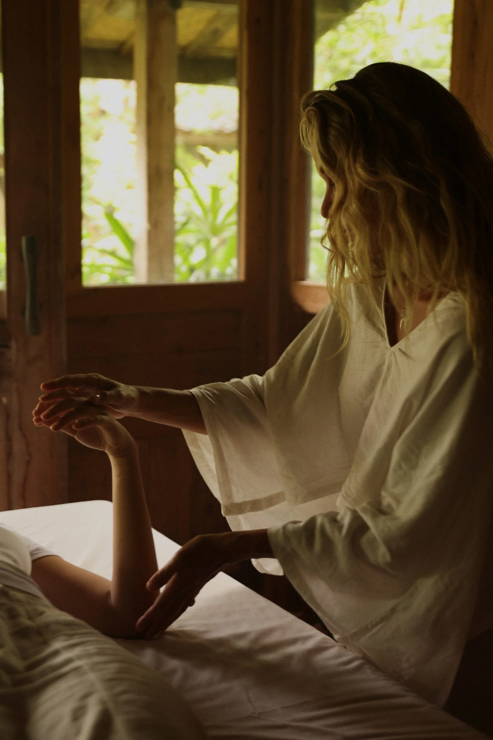 Une femme avec des cheveux blonds bouclés offre un massage à une personne allongée sur un lit dans une pièce en bois avec une lumière naturelle