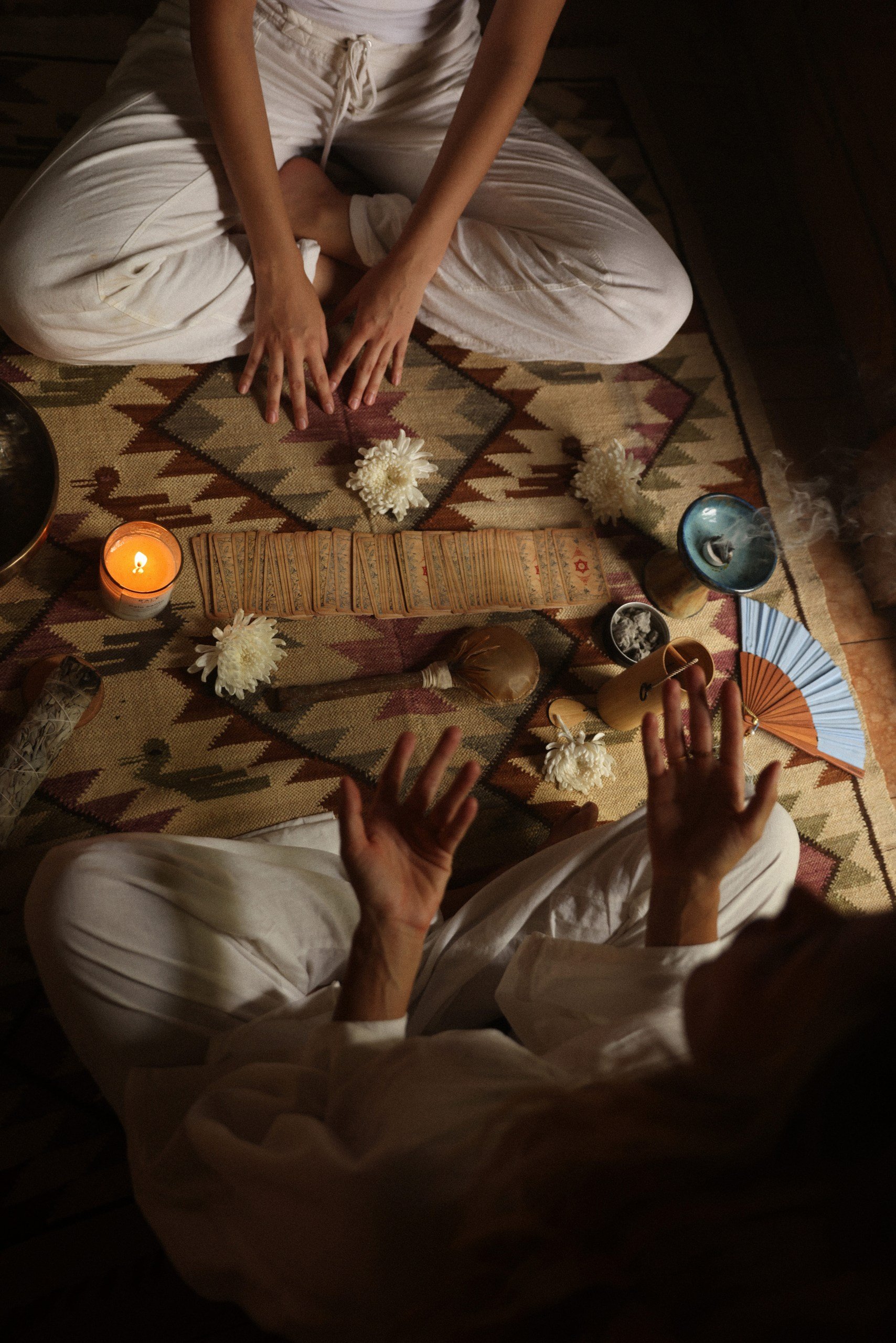 Deux personnes assises en tailleur sur un tapis, entourées de tarots, bougies, fleurs, fumée d'encens, et un éventail, dans une ambiance mystérieuse et chaleureuse.