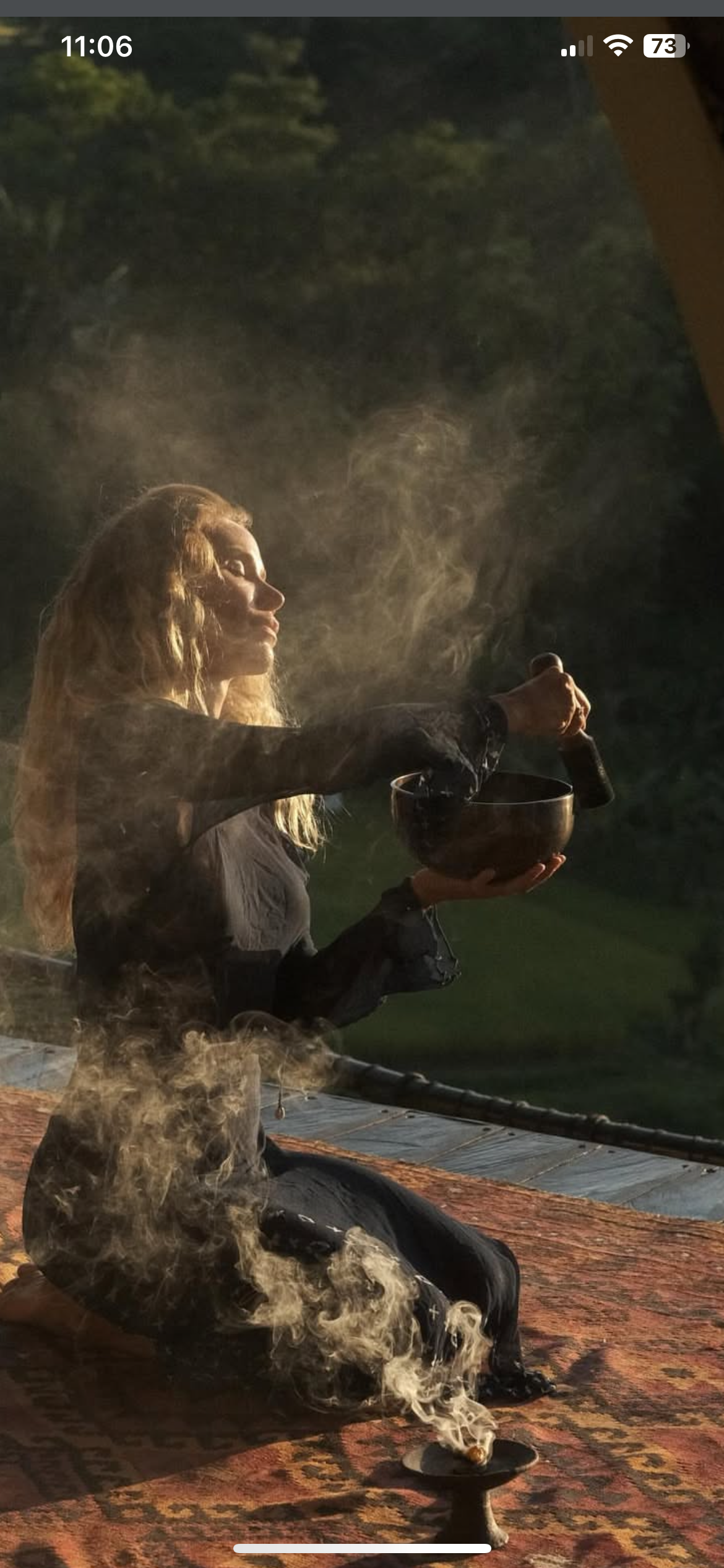 Une femme avec des cheveux blonds longs, vêtue de noir, assise sur un tapis moquette, tenant une coupe en bois dans une main et un marteau en pierre dans l'autre, avec de la fumée qui s'élève dans l'air, dans un environnement extérieur avec arbres en arrière-plan.