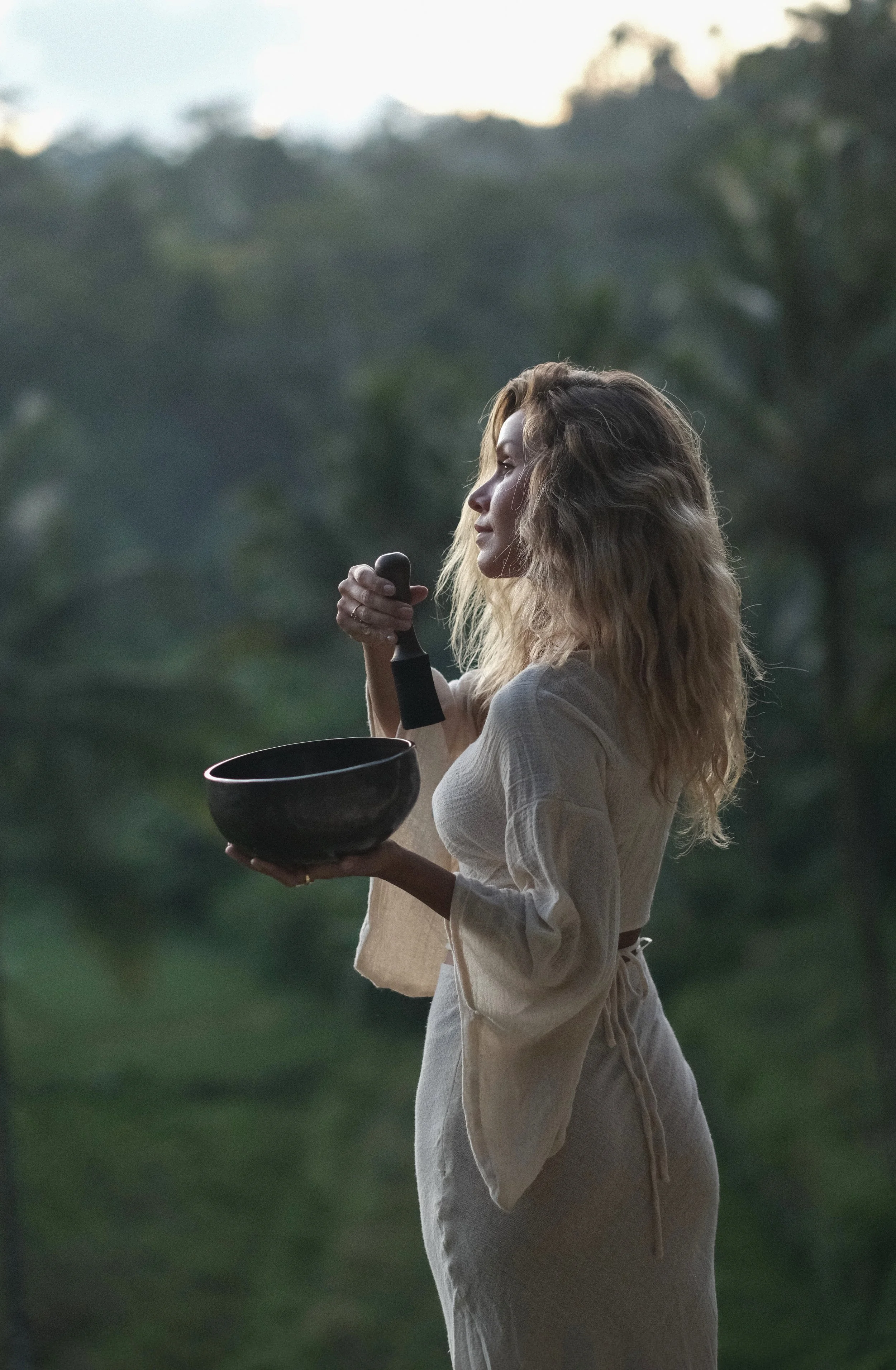 Femme avec des cheveux bouclés, portant une robe beige, tenant une coupe en bois et un bâton de percussion en bois dans un environnement naturel.