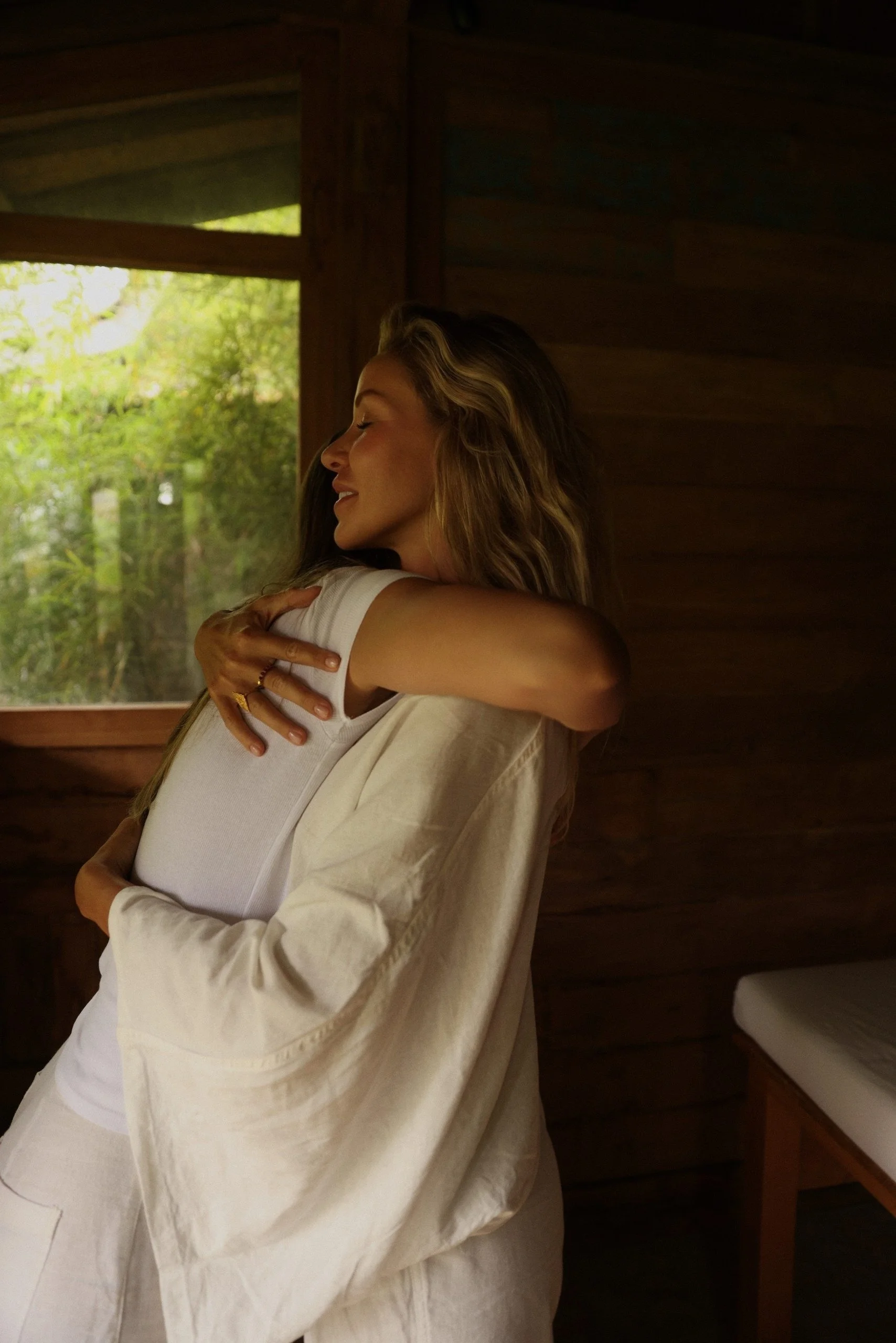 Deux femmes s'embrassent dans une pièce en bois avec une fenêtre montrant des arbres verts à l'extérieur.