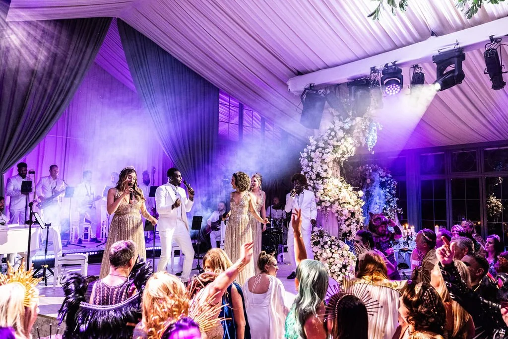 Group of performers singing on stage at a wedding reception with an audience dancing and celebrating.
