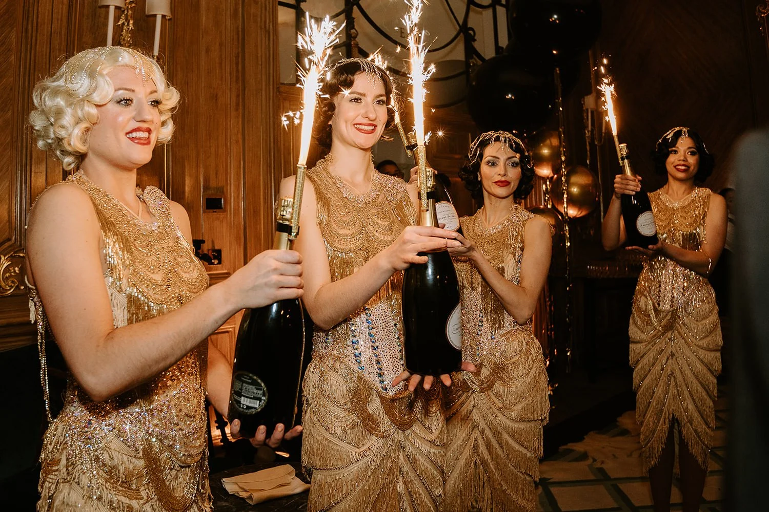 Four women dressed in 1920s-style gold bead and fringe dresses holding bottles with sparklers at a celebration or party.