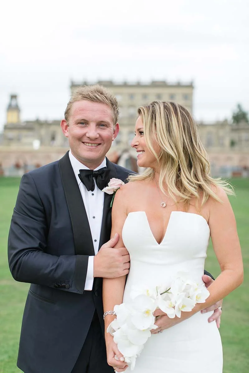 A newlywed couple on their wedding day outdoors, with the groom in a black tuxedo and the bride in a white strapless wedding gown holding a bouquet of white flowers, standing in front of a historical building.
