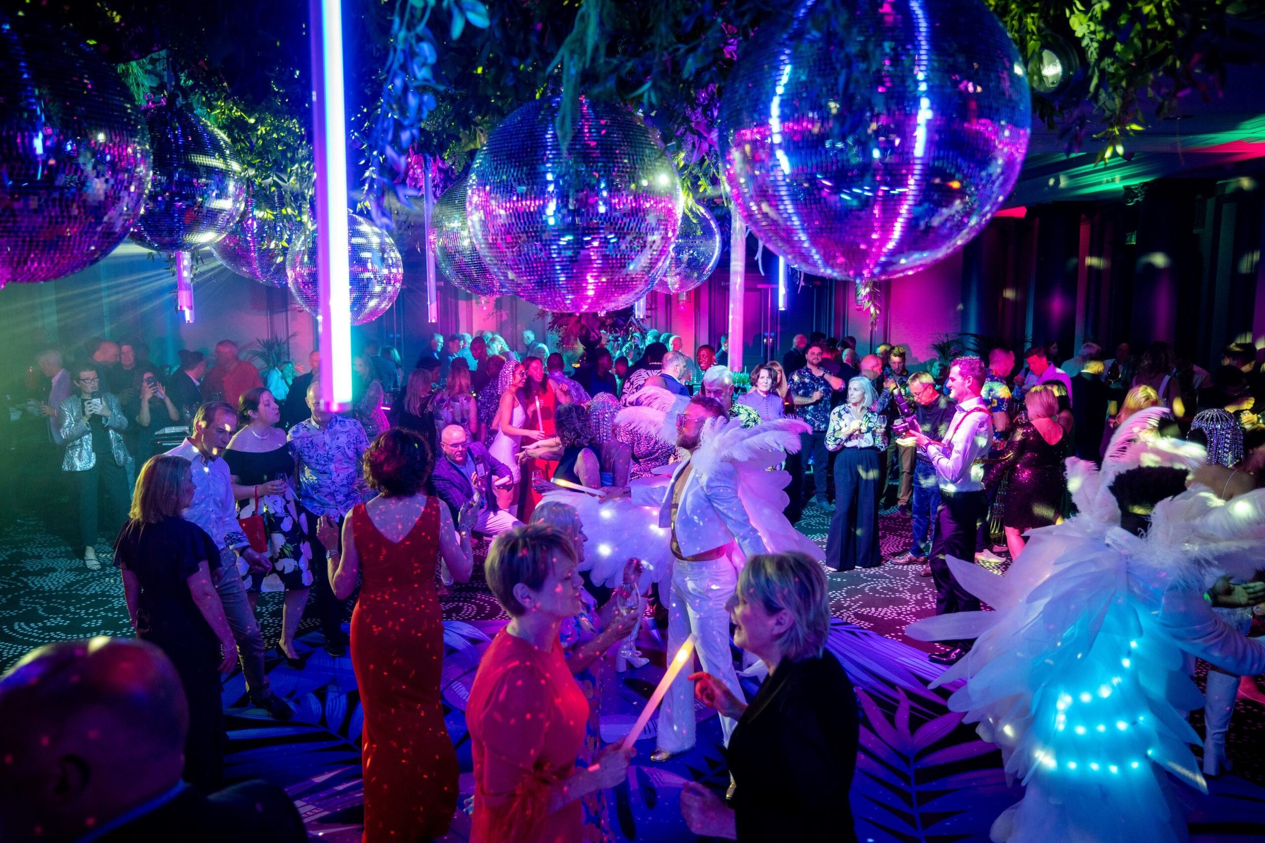 Vibrant party scene with many people dancing and socializing under large disco balls with colorful lighting.