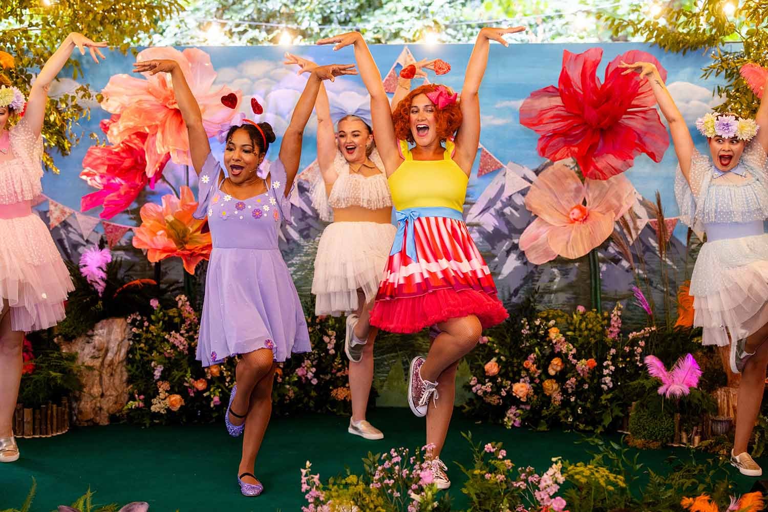 Women and girls dancing on stage with a colorful garden and mountain backdrop, wearing floral and pastel skirts and tops.