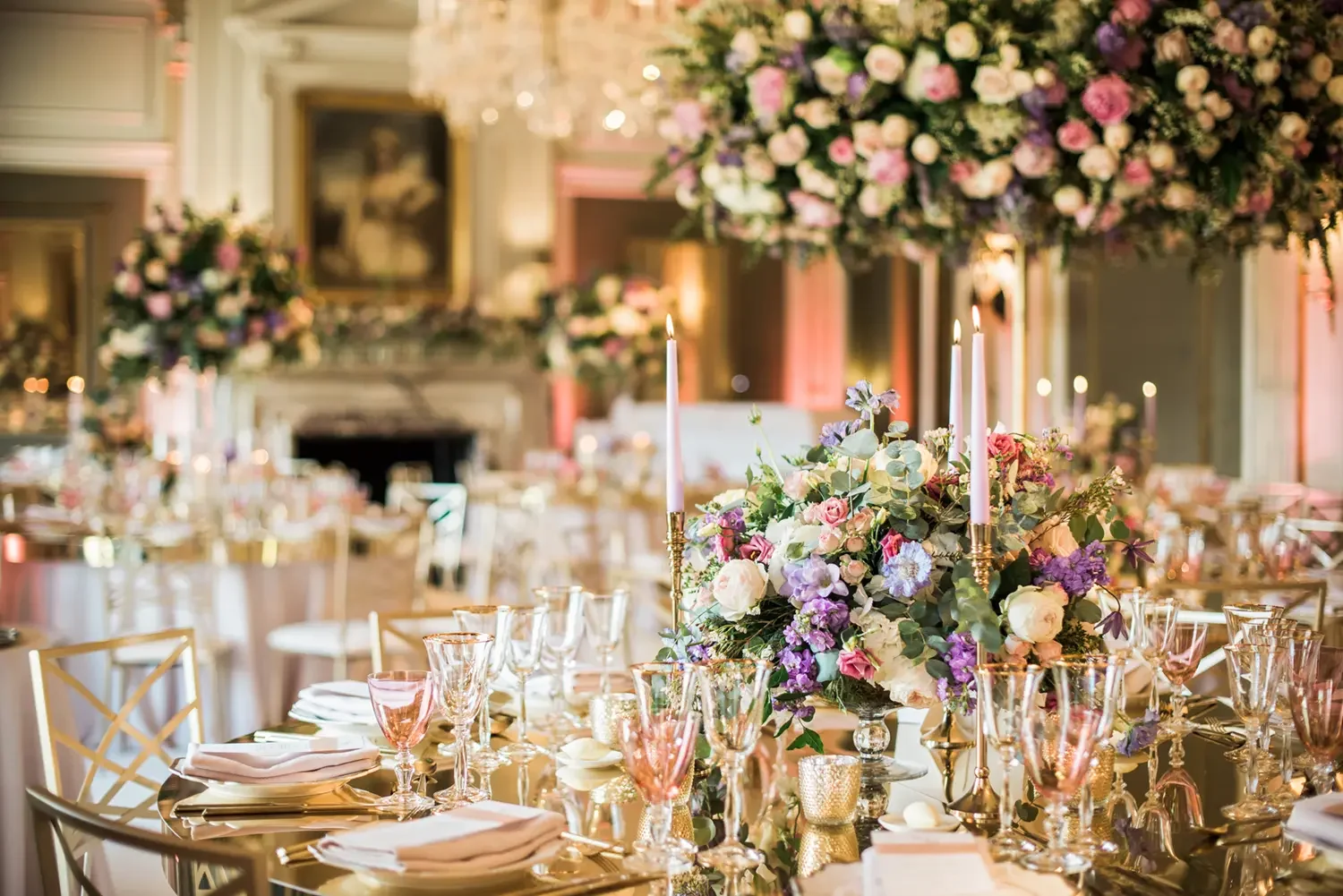 Elegant banquet table decorated with a large floral centerpiece, tall candles, pink and purple glassware, and gold accents in a luxurious reception hall.