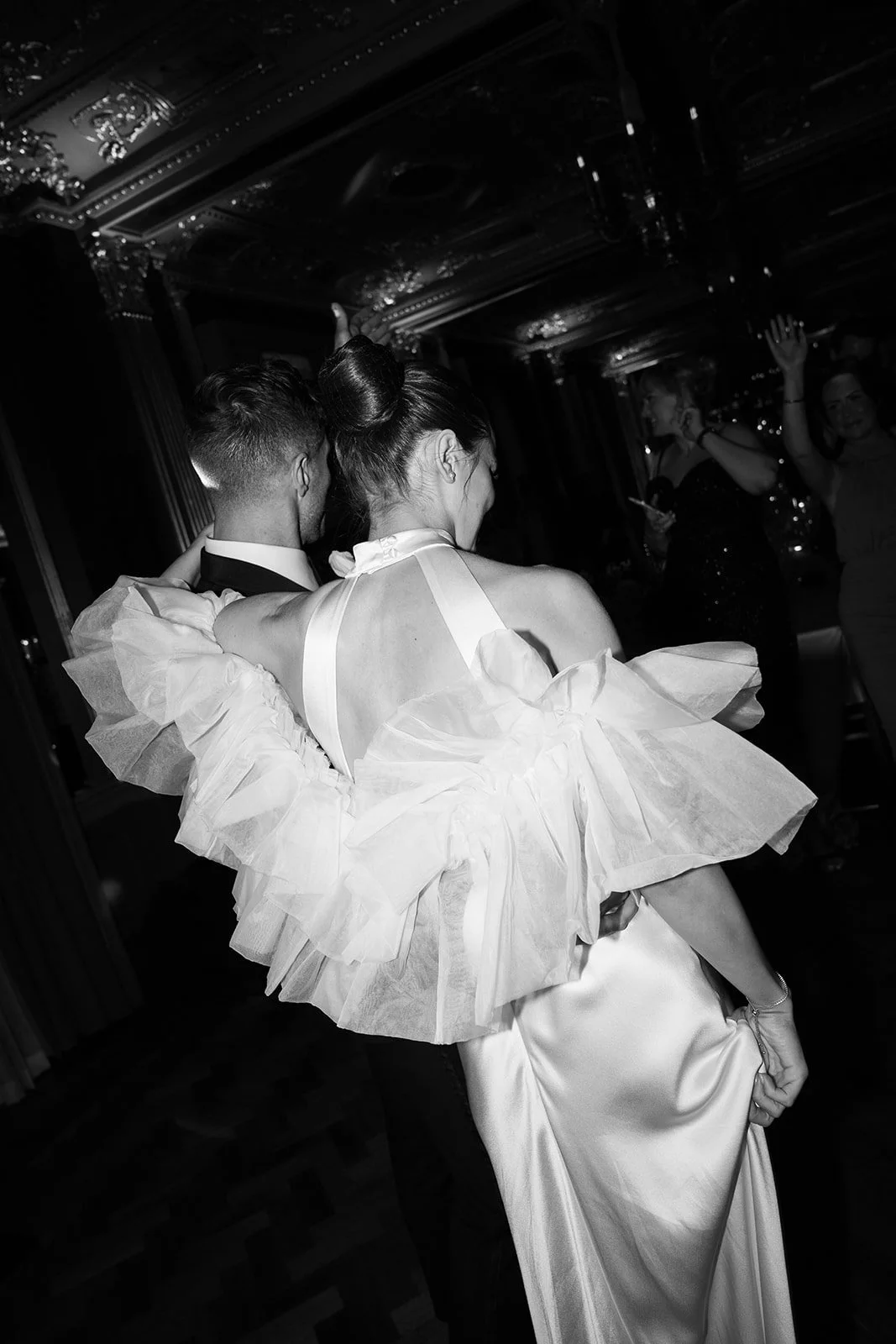 A couple dancing at a formal event, with women in elegant dresses in the background. The woman is wearing a flowing gown with ruffles and a large bow on the back, and the man is in a tuxedo. The scene is indoors with ornate ceiling details.