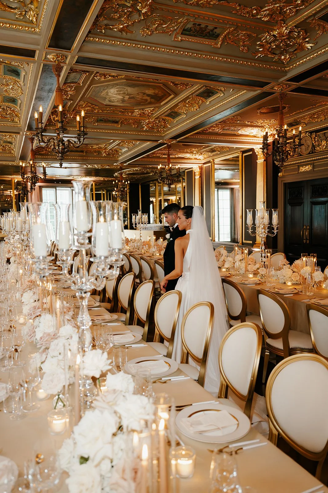 A bride and groom standing at a long, elegantly decorated wedding reception table in a luxurious, ornate room with gold accents and chandeliers.