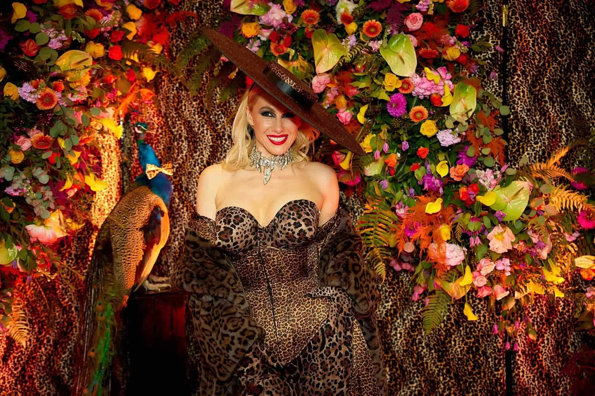 A woman in a leopard print dress and large brim hat smiling, standing in front of a vibrant background with leopard print fabric and colorful flowers, with a large blue and orange parrot beside her.