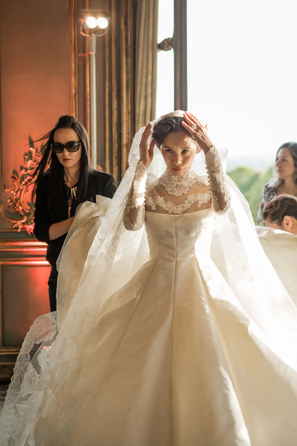A bride in a lace wedding dress adjusting her veil, with her wedding gown flowing outwards, inside a decorated room with large windows and curtains, accompanied by two women, one wearing sunglasses.