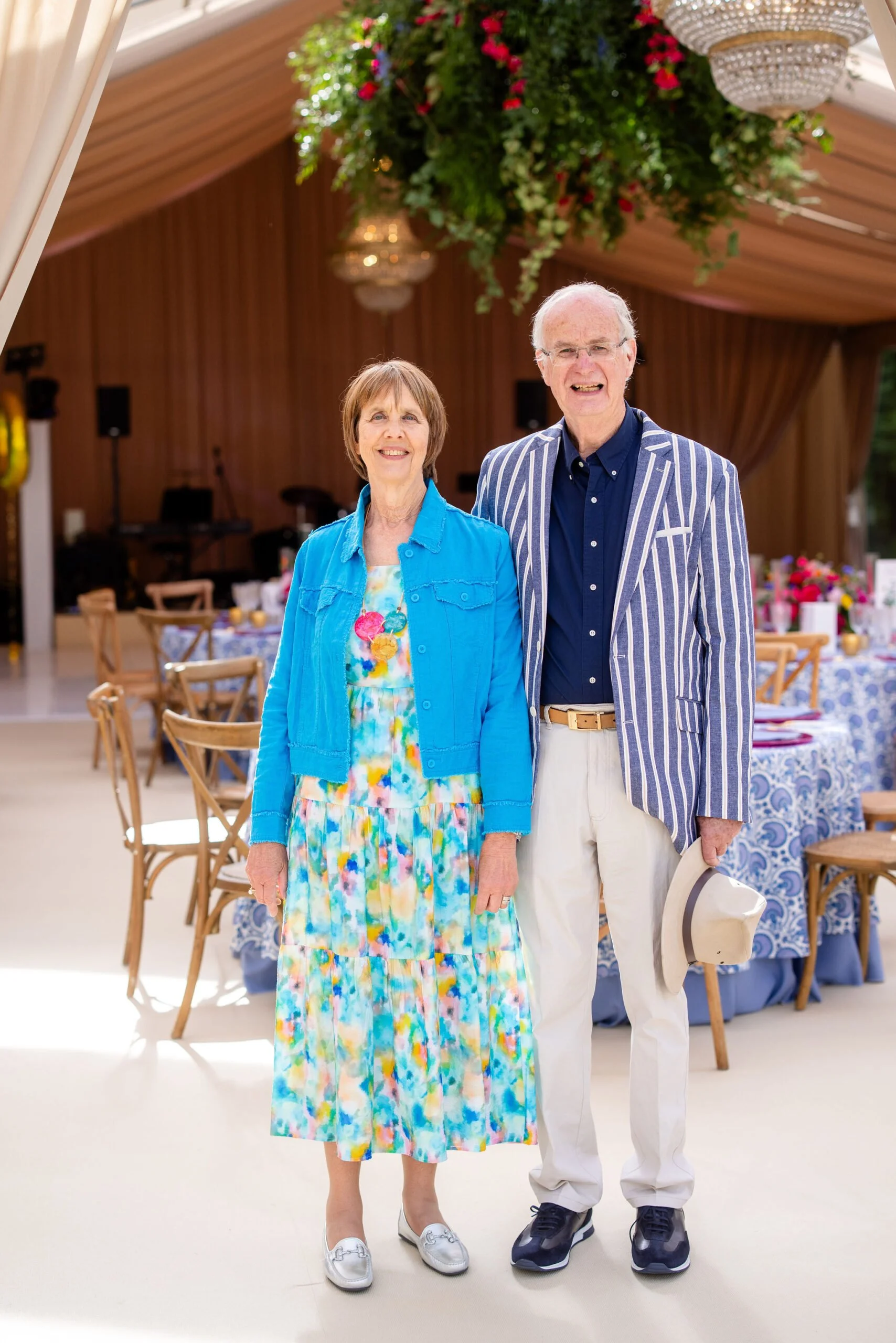 An elderly couple stands indoors at a decorated event space with tables, chairs, and a chandelier overhead. They are both smiling, dressed in colorful, coordinated summer outfits, and the man is holding a hat.