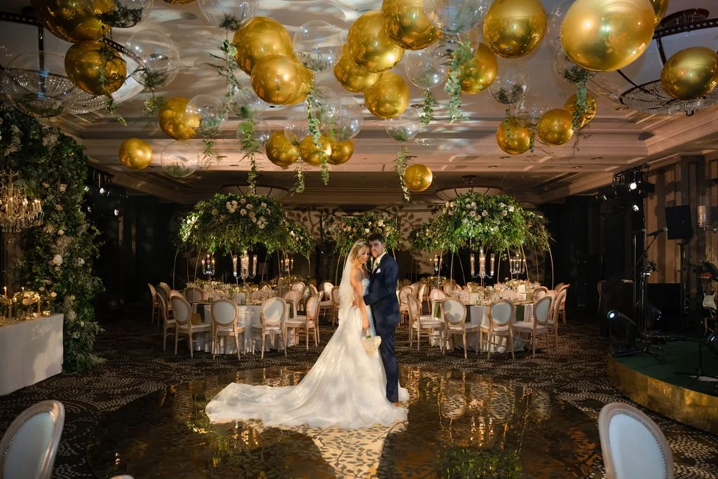 A bride and groom celebrating their wedding in a decorated ballroom with gold and transparent balloons hanging from the ceiling, large floral arrangements on the tables, and elegant table settings.
