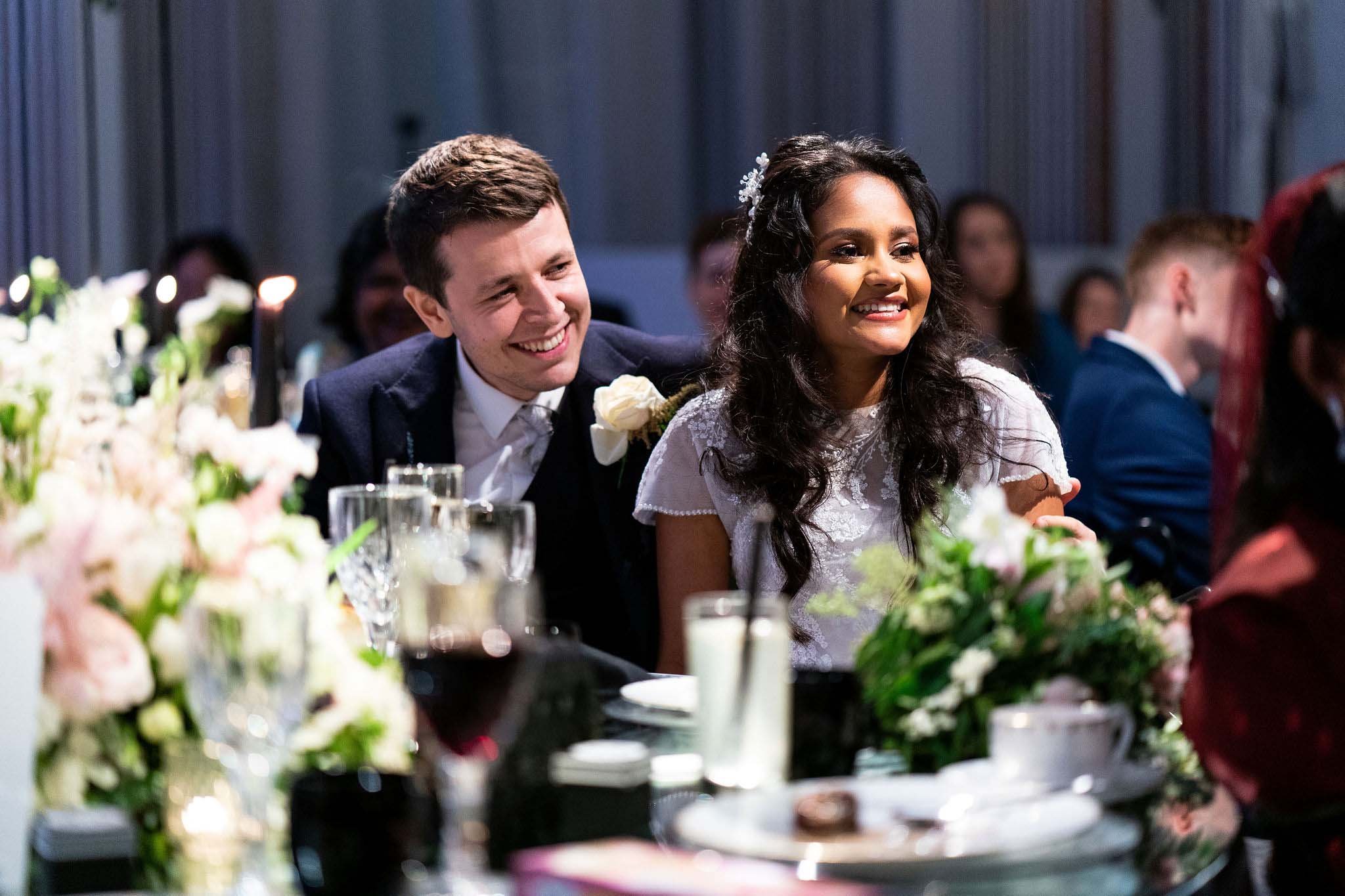 A wedding reception with a bride and groom sitting at a table, smiling, surrounded by floral arrangements, glasses, and candles.