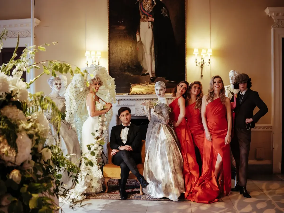 A group of women in elegant dresses and two men, one in a tuxedo and one in a suit with a hat, posing in an ornate room with a large portrait painting and a fireplace, surrounded by decorative furniture and flowers.