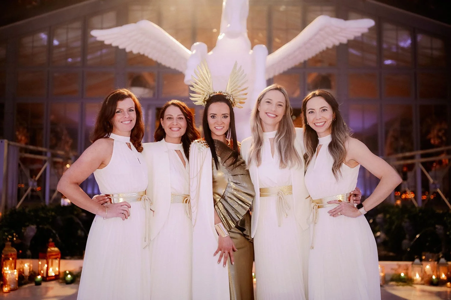 Five women dressed in white, with the woman in the center wearing gold, standing in front of a large angel sculpture with wings and a halo, inside a building with glass walls and warm lighting.