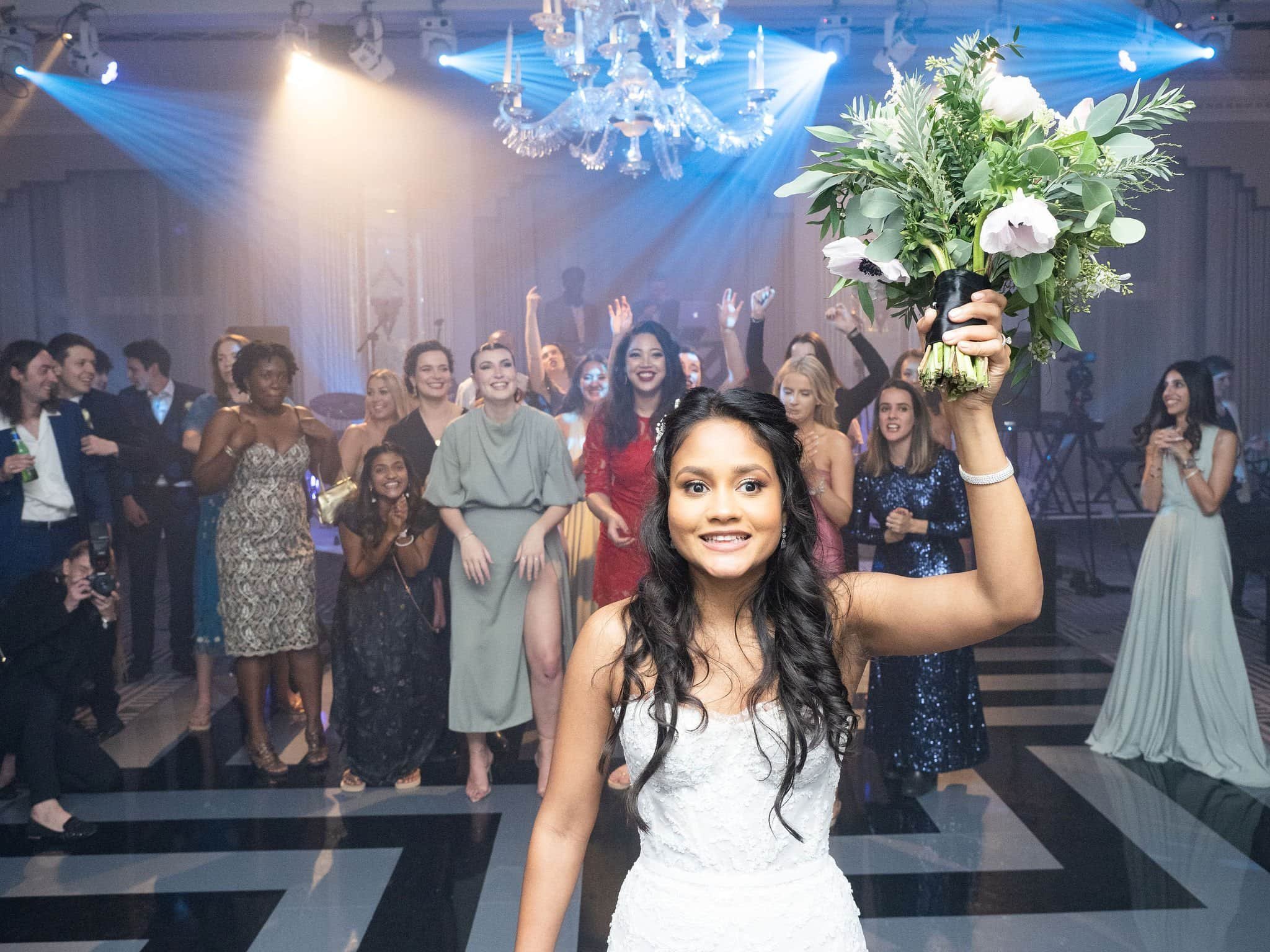 A bride holding a bouquet of flowers at her wedding reception with guests in the background dancing and celebrating.