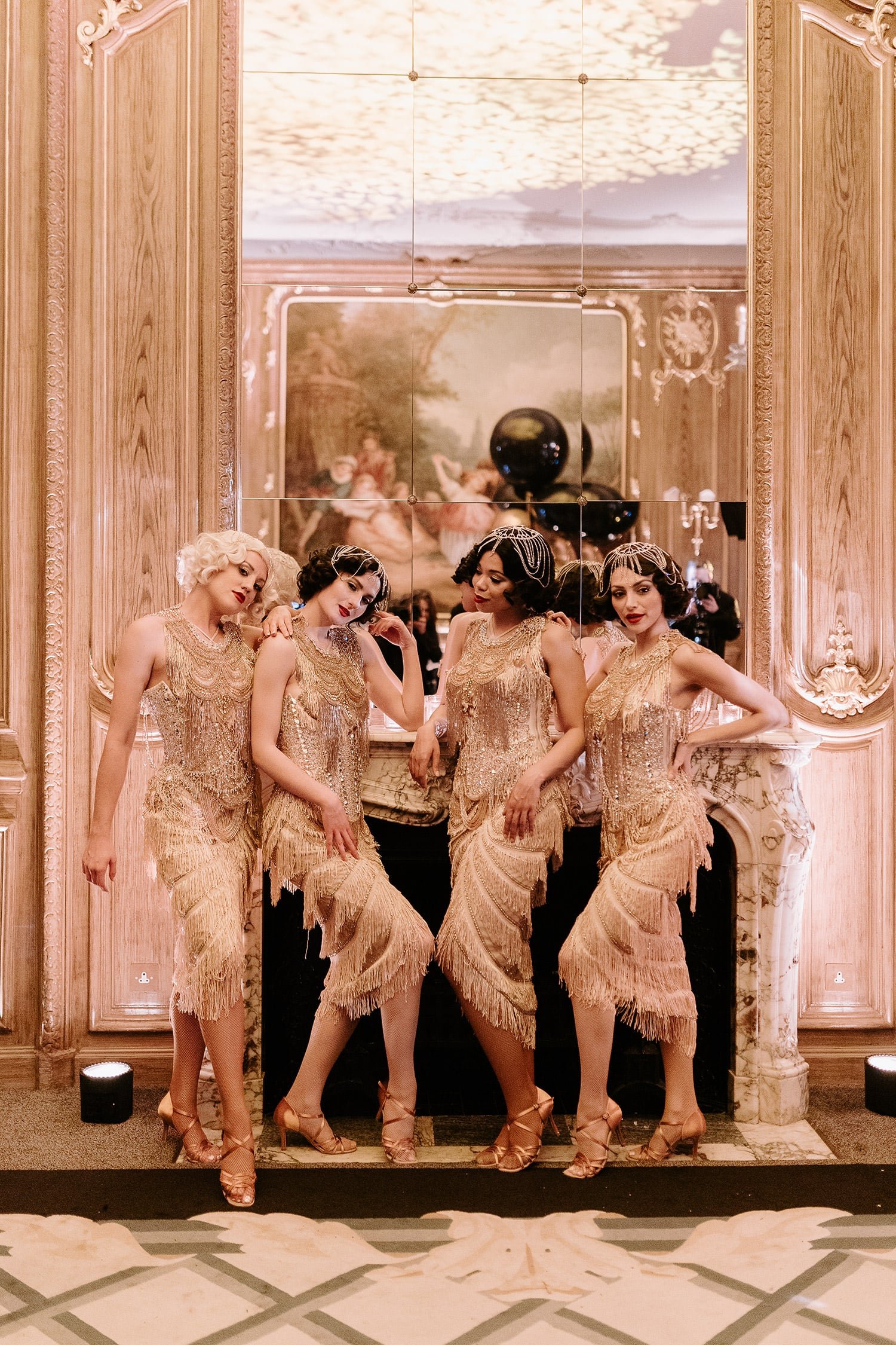Four women dressed in vintage, flapper-style gold dresses and heels posing inside an ornate room with a large mirror and a fireplace.