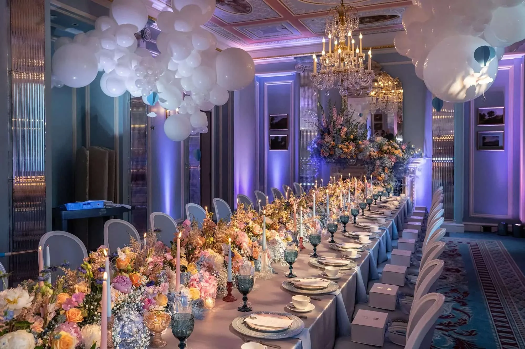 Elegant banquet table with floral centerpieces, candles, and tableware in a decorated event hall with balloons hanging from the ceiling and chandeliers.